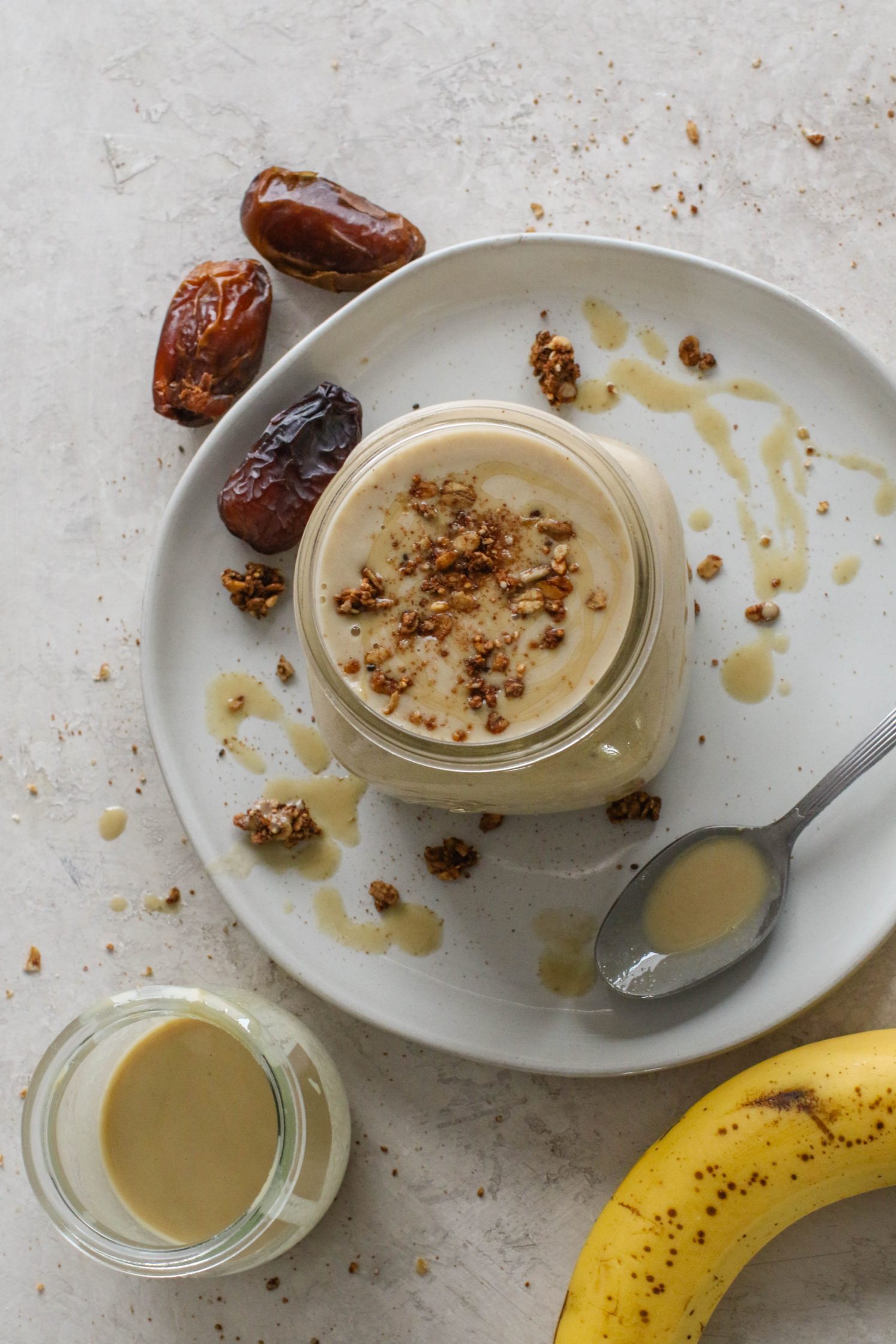 Tahini Date Shake served in a mason jar on a plate topped with granola and tahini with dates and banana on the side by Flora & Vino