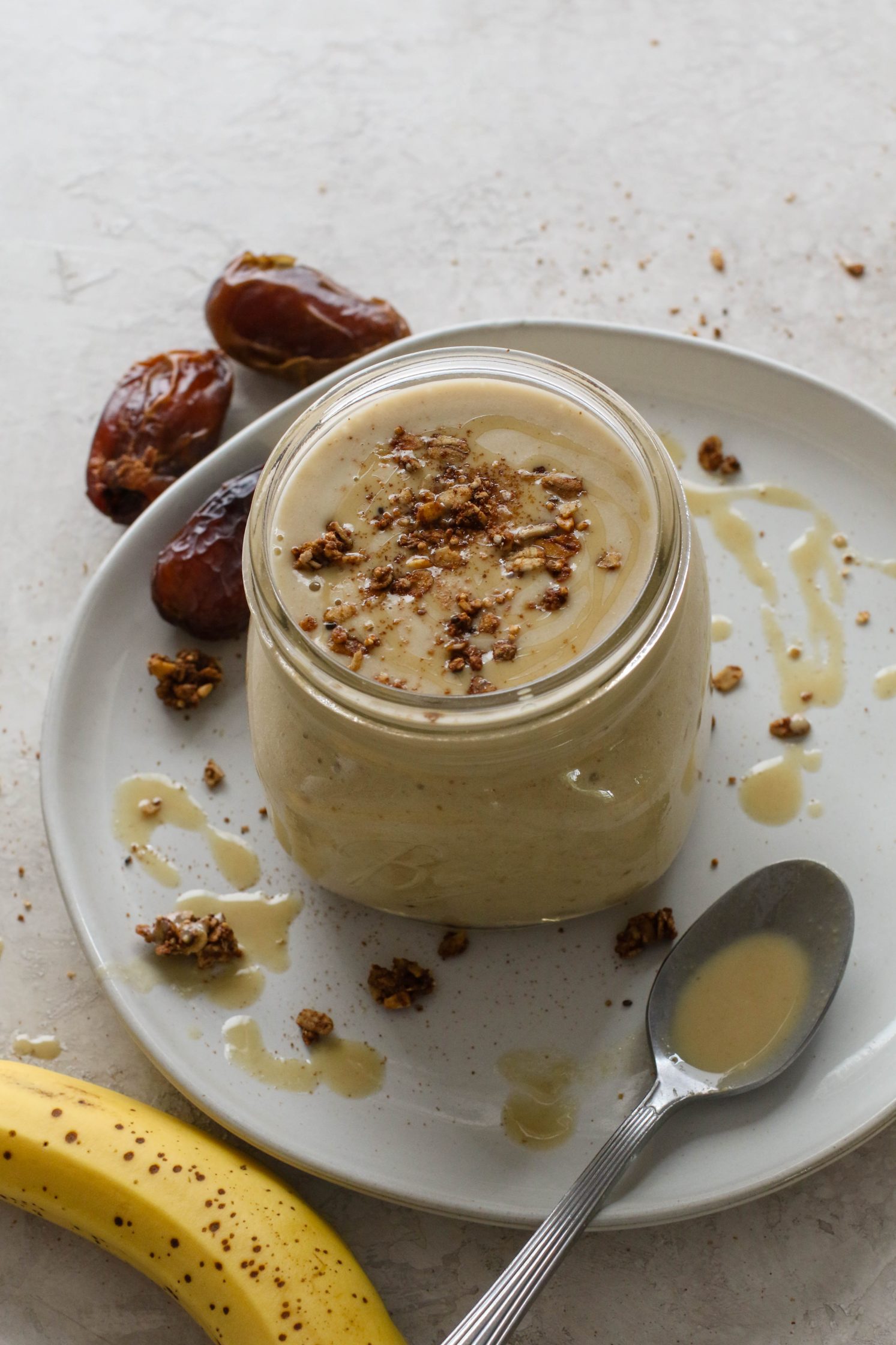 Tahini Date Shake served in a mason jar on a plate topped with granola and tahini with dates and banana on the side by Flora & Vino