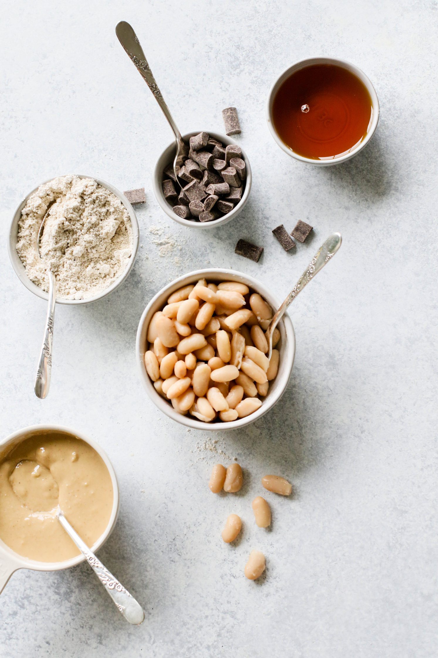 white beans in bowl with a spoon, maple syrup in bowl, chocolate chunks in bowl with a spoon, oat flour in a bowl with a spoon, and tahini in bowl with spoon by Flora & Vino