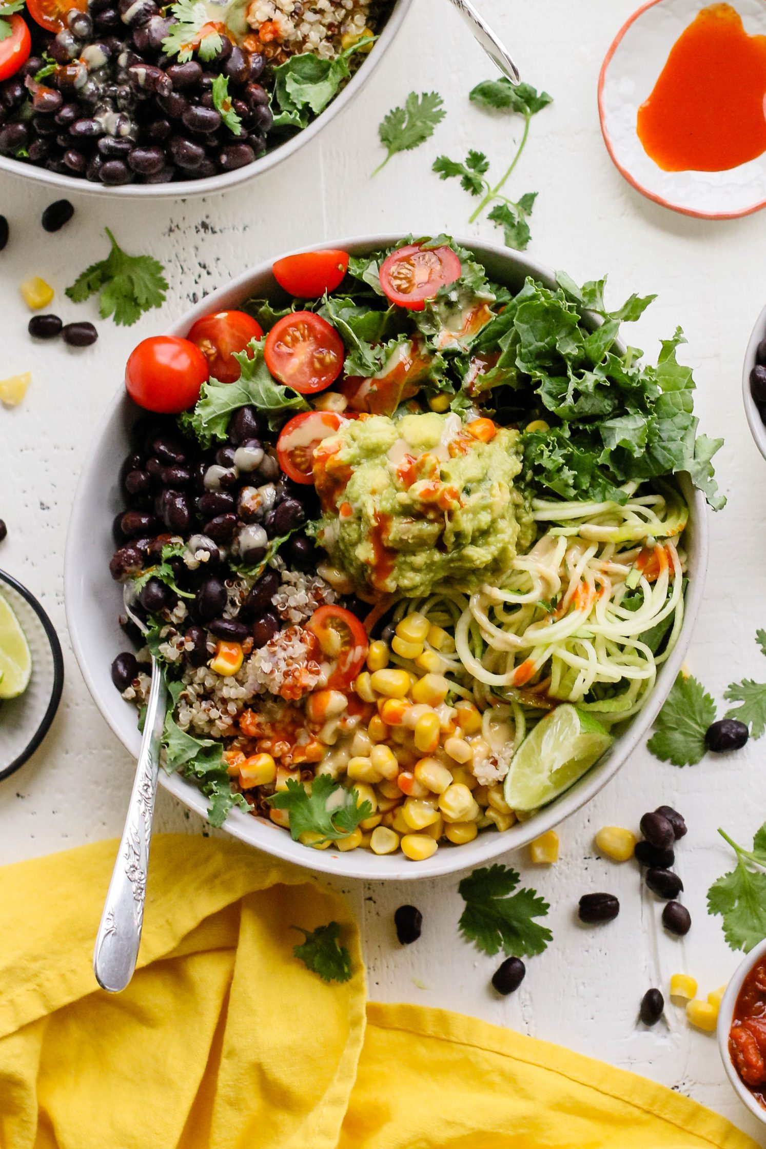 Easy Mexican Macro Bowls served in white bowl with hot sauce, guacamole, corn, cherry tomatoes, kale, zucchini spirals, black beans, and lime with a fork