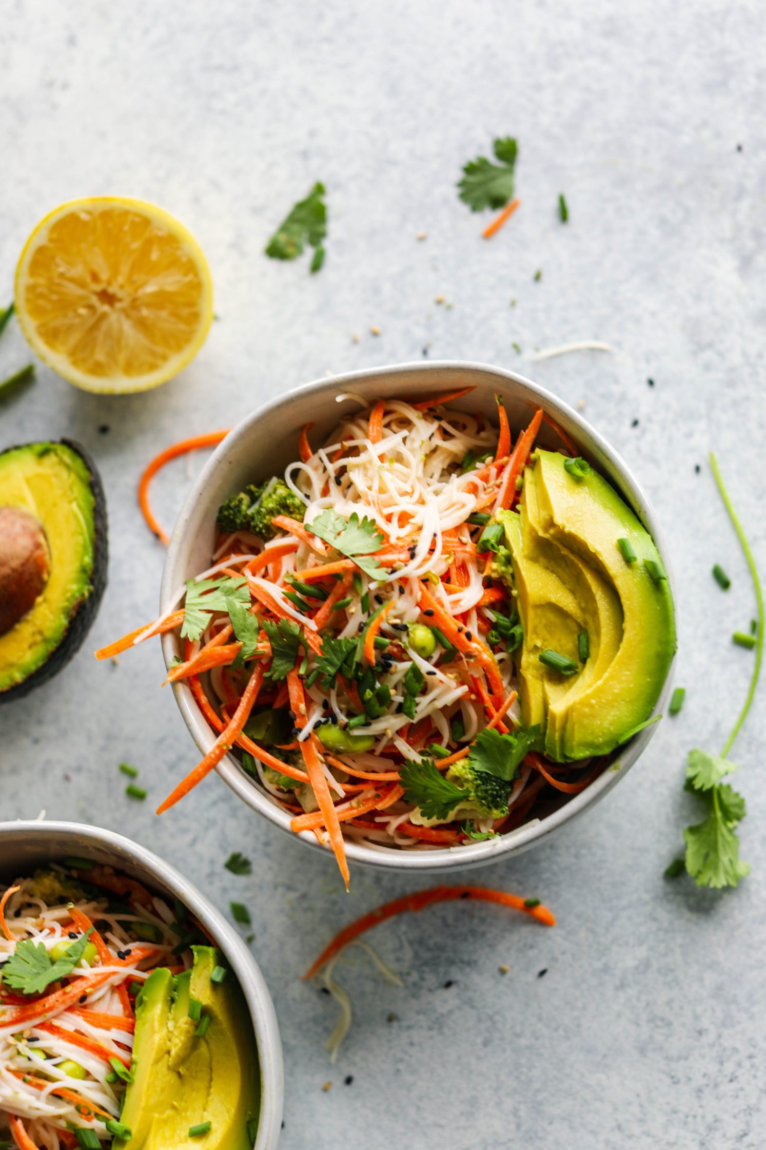 Rainbow Rice Noodle Bowls served in bowl topped with avocado, sesame seeds, and cilantro with two forks