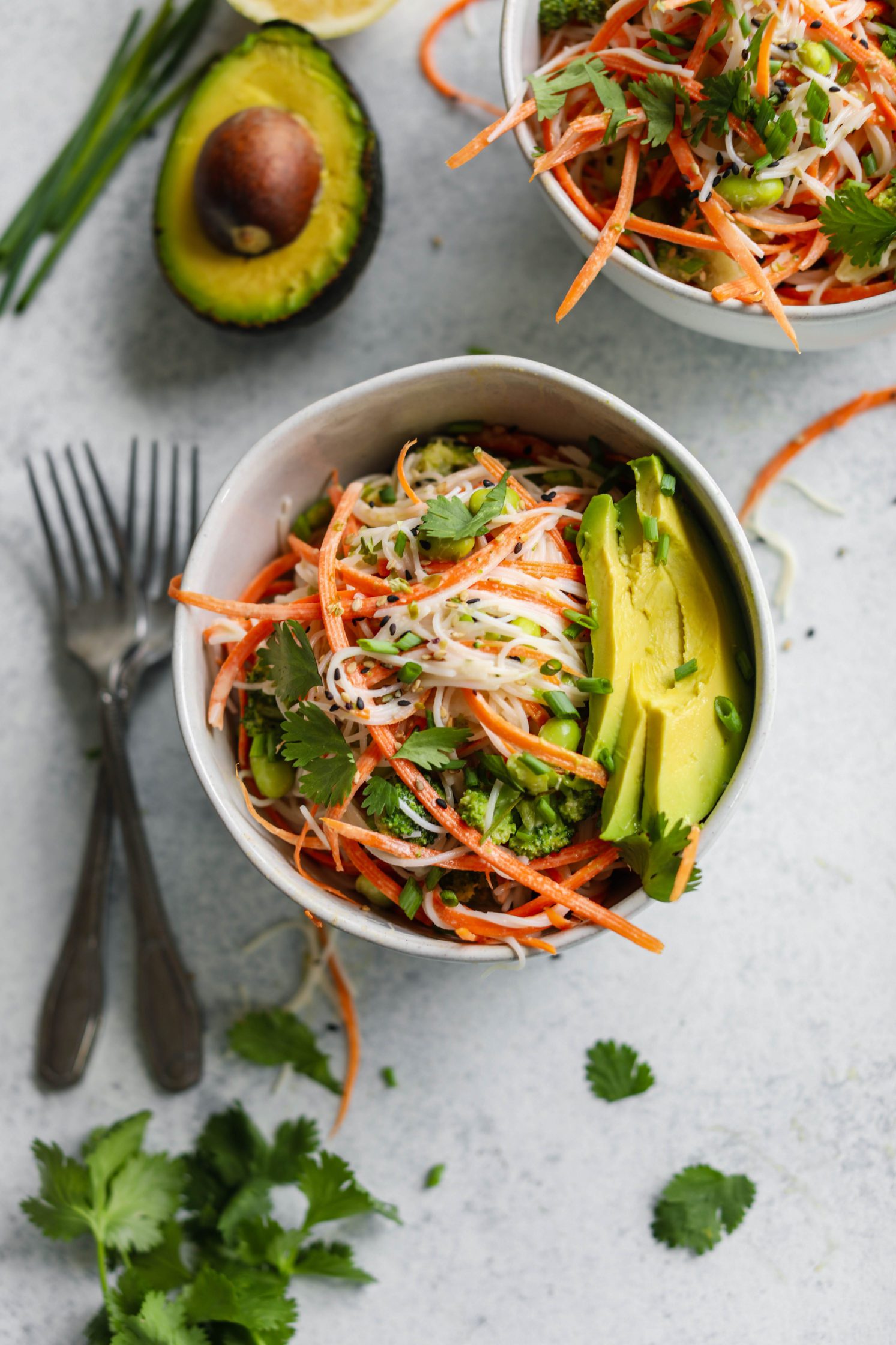 Rainbow Rice Noodle Bowls served in bowl topped with avocado, sesame seeds, and cilantro with two forks