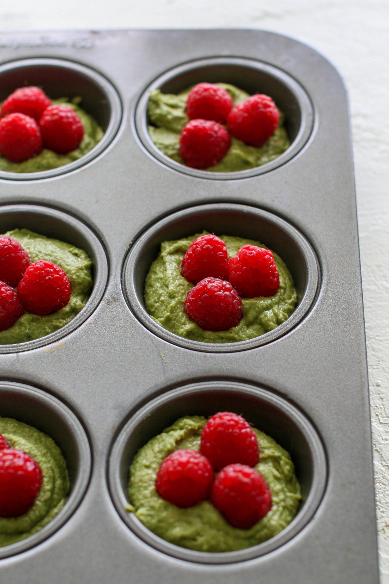 matcha muffin batter in muffin tins with raspberries on top by Flora & Vino