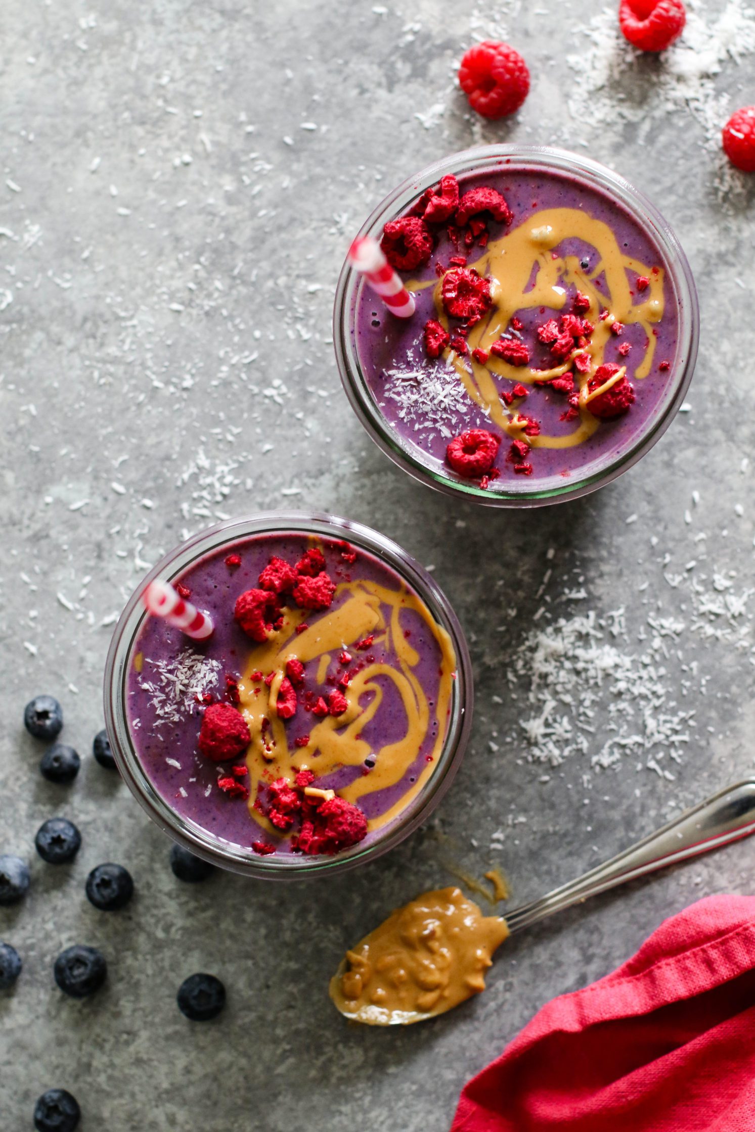 Peanut Butter Jelly & Beet Shakes served in mason jars topped with peanut butter and freeze-dried raspberries with straws. Spoon of peanut butter on the side with a red towel and scattered berries.