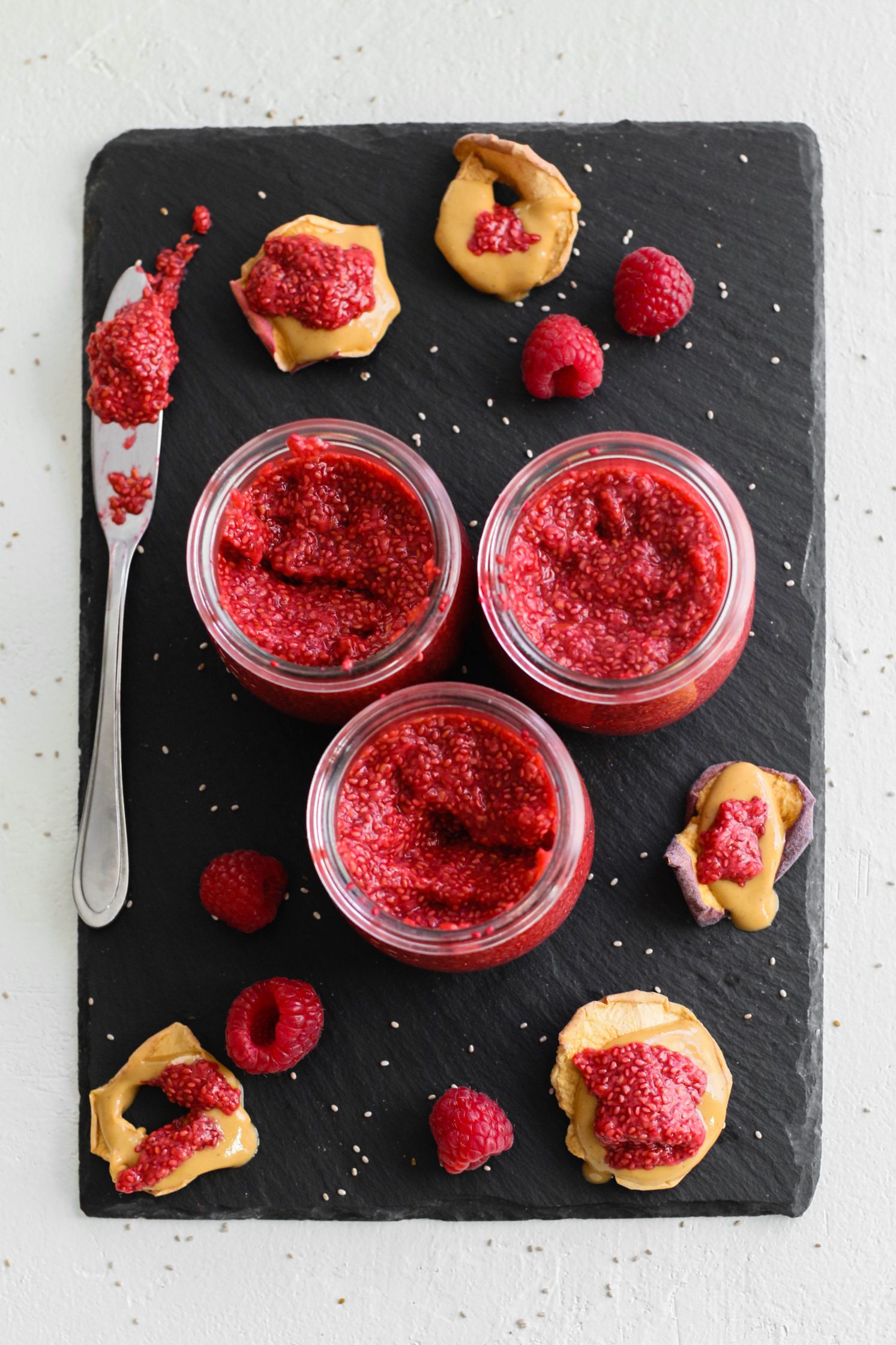 No-Sugar-Added Raspberry Chia Seed Jam in small jars with raspberries scattered and apple chips topped with peanut butter and jam