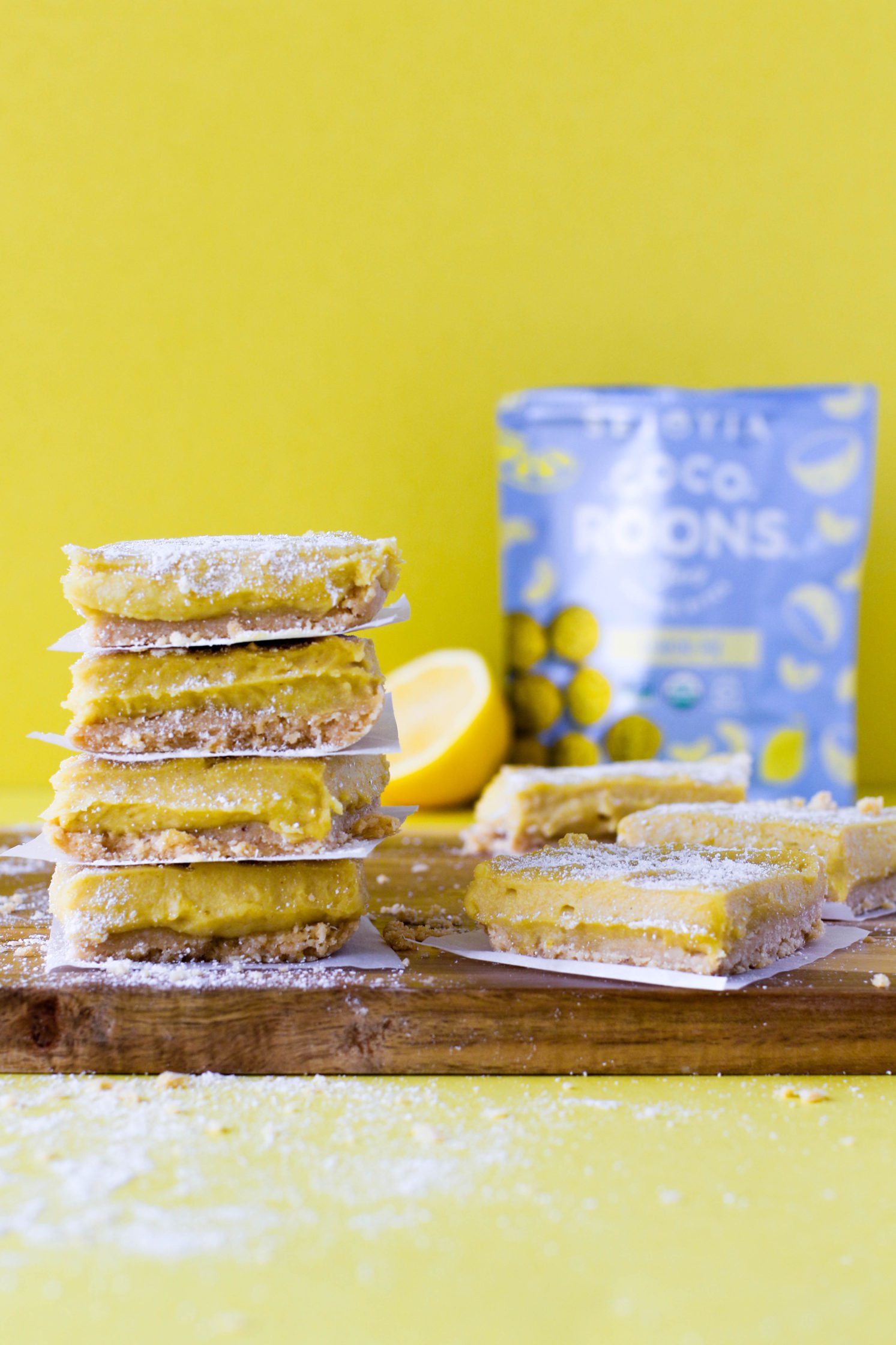 No-Bake Lemon Pie Bars stacked on wooden board with yellow background topped with coconut flour with lemons