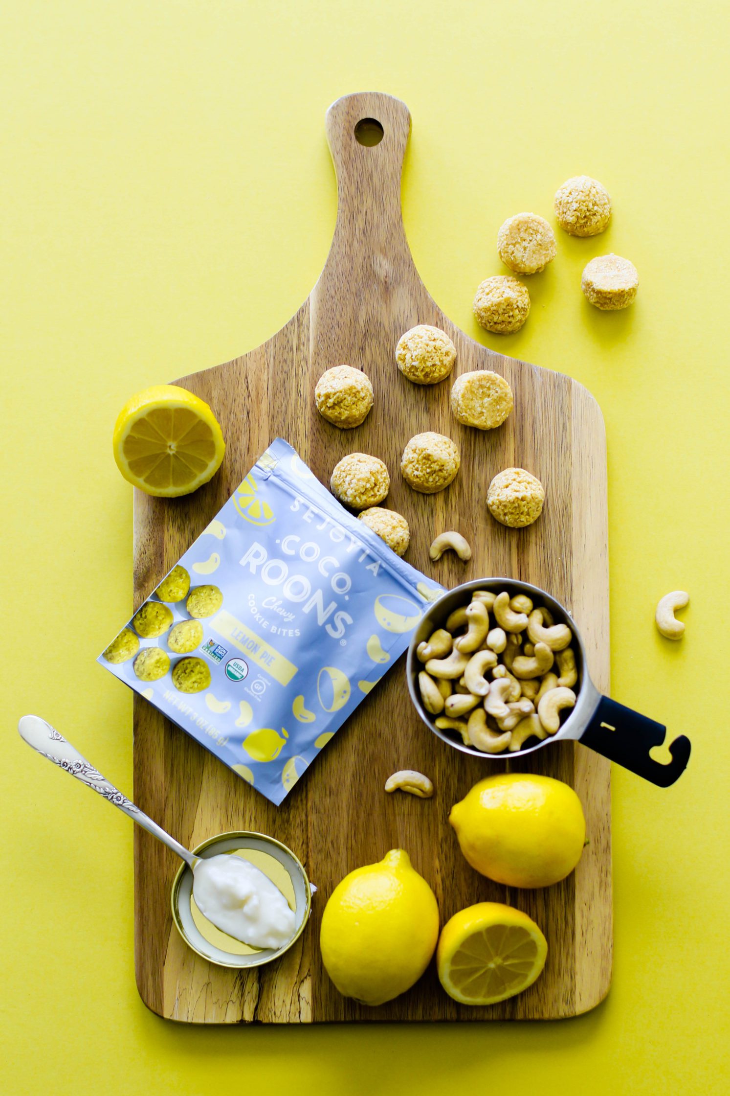 Coco-Roons spilling out of bag on wooden board with cup of cashews and fresh lemons with spoon of coconut butter on yellow background