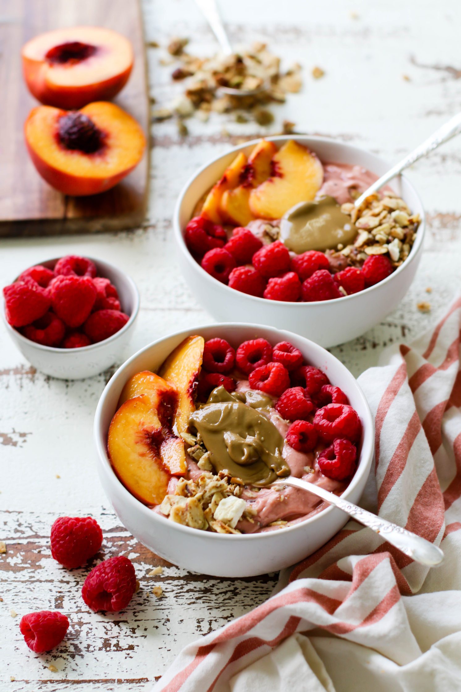 Peach Raspberry Sunbutter Smoothie Bowls served with spoons of SunButter, fresh raspberries, sliced peaches, and granola on distressed white board with pink and white striped towel
