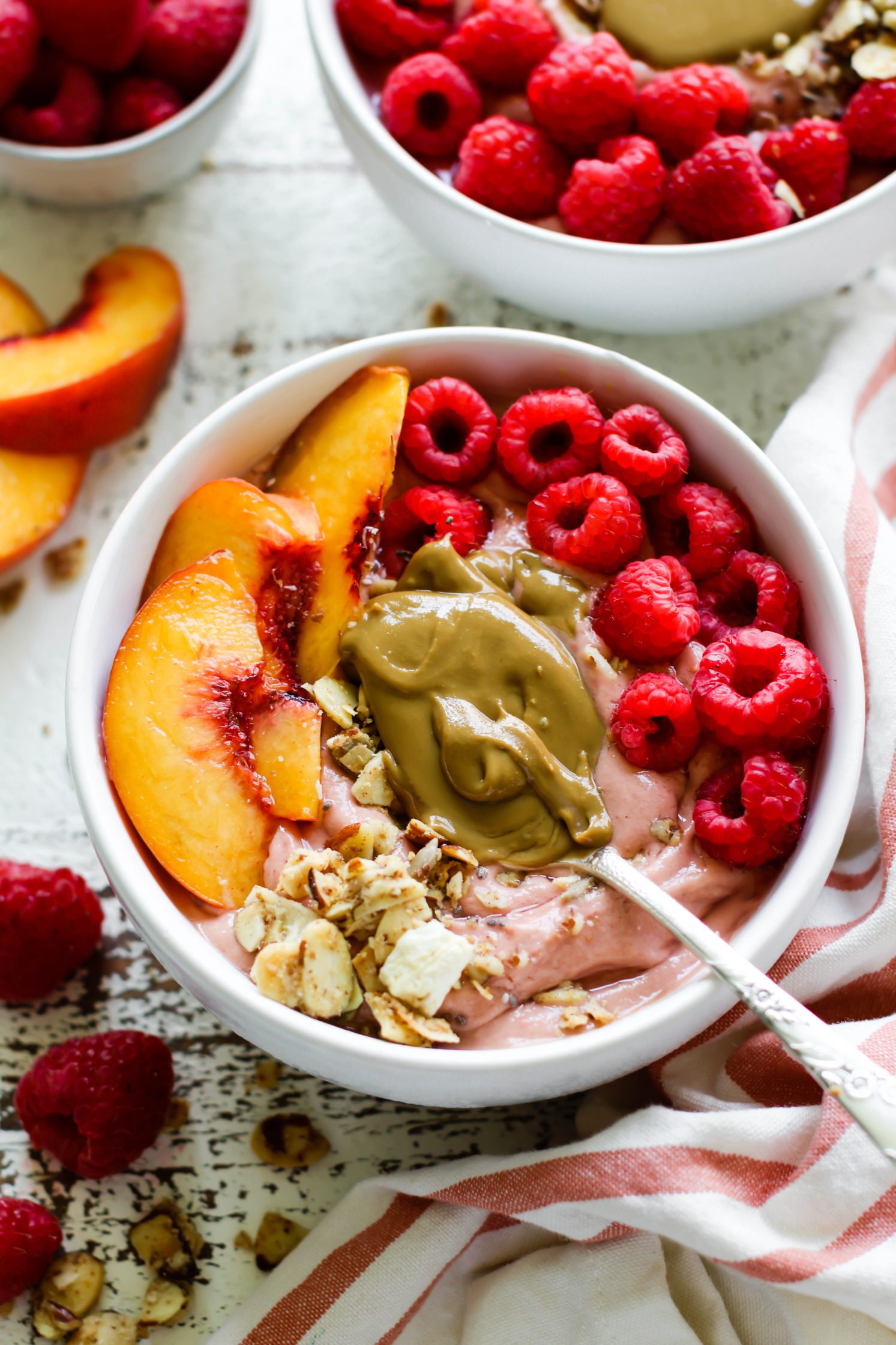 Peach Raspberry Sunbutter Smoothie Bowls served with spoons of SunButter, fresh raspberries, sliced peaches, and granola on distressed white board with pink and white striped towel
