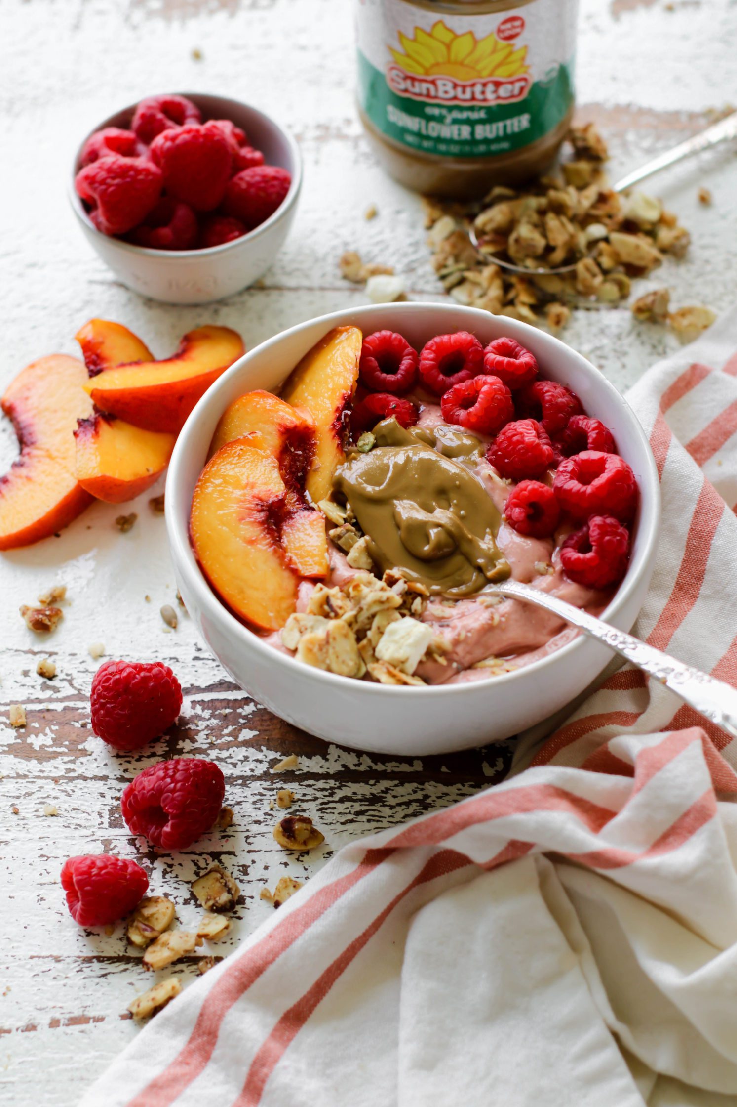 Peach Raspberry Sunbutter Smoothie Bowls served with spoons of SunButter, fresh raspberries, sliced peaches, and granola on distressed white board with pink and white striped towel