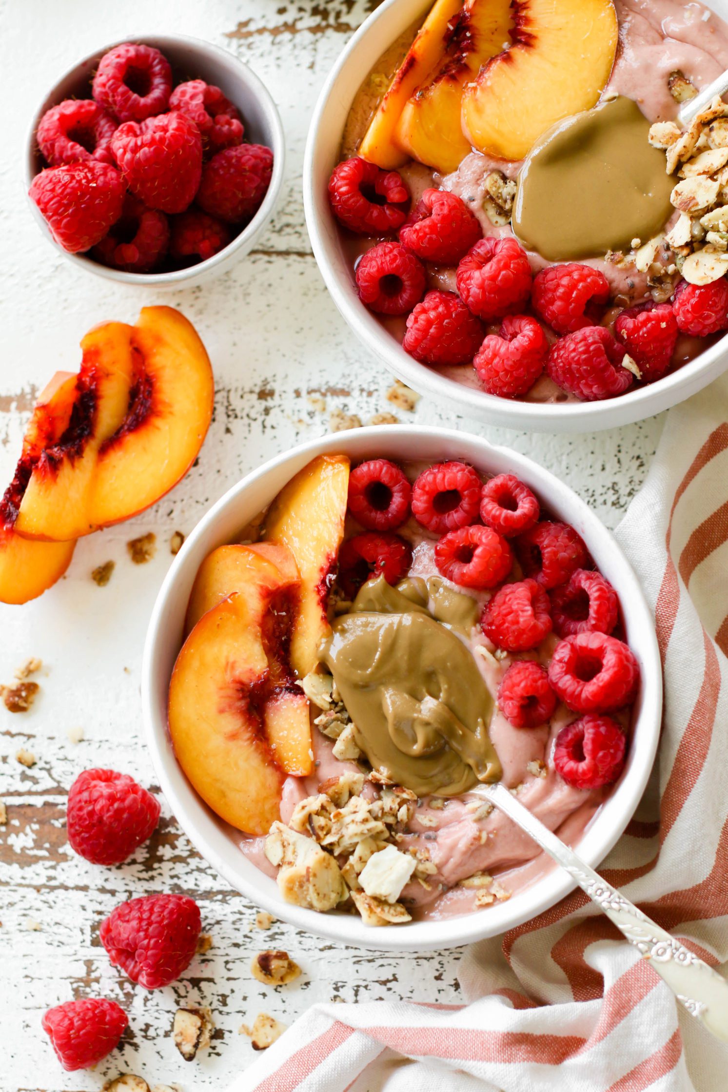 Peach Raspberry Sunbutter Smoothie Bowls served with spoons of SunButter, fresh raspberries, sliced peaches, and granola on distressed white board with pink and white striped towel