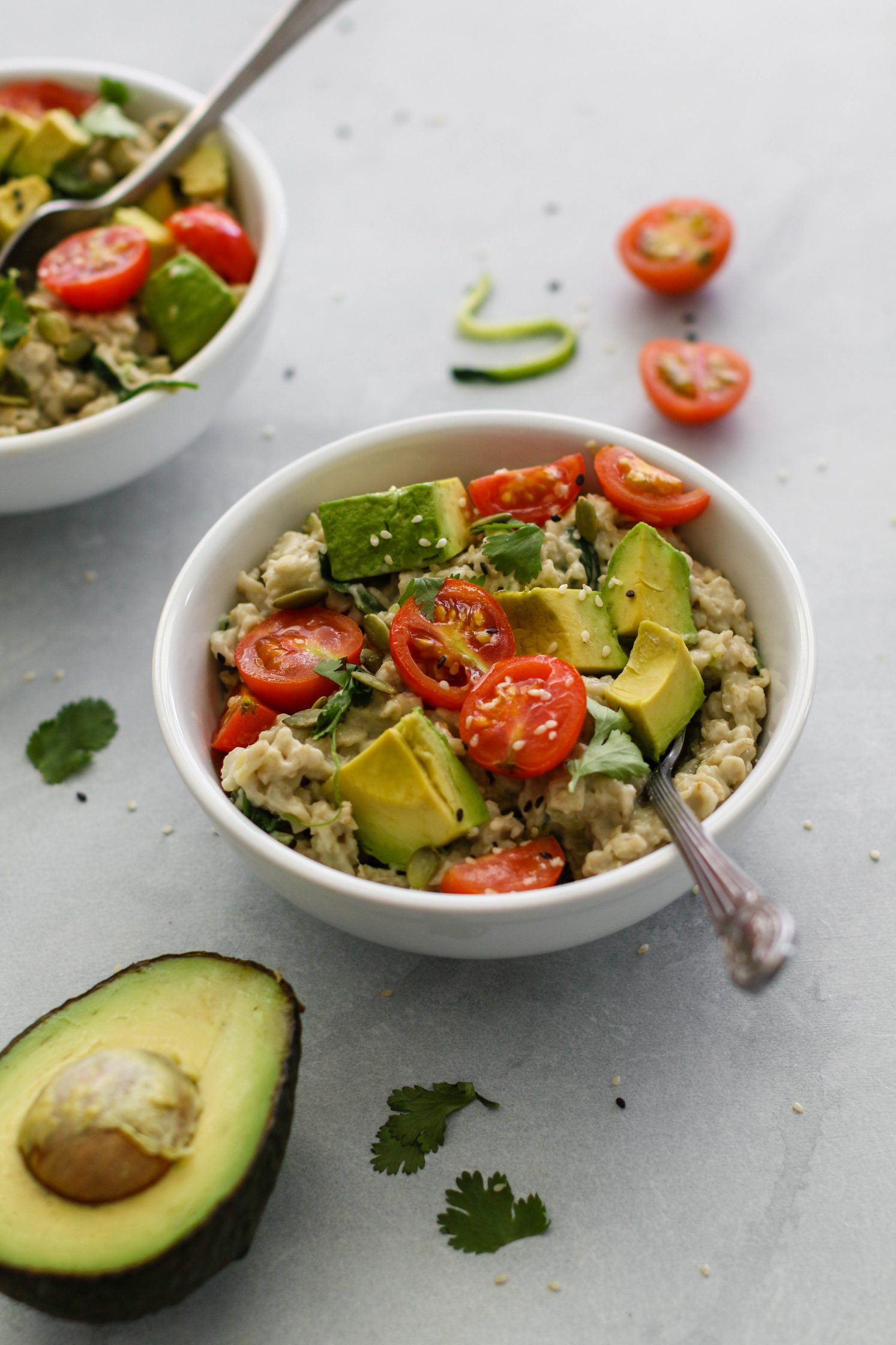Savory Zucchini Oats (Zoats) served in bowls topped with cherry tomatoes and avocado with spoons surrounded b  avocado and cherry tomatoes