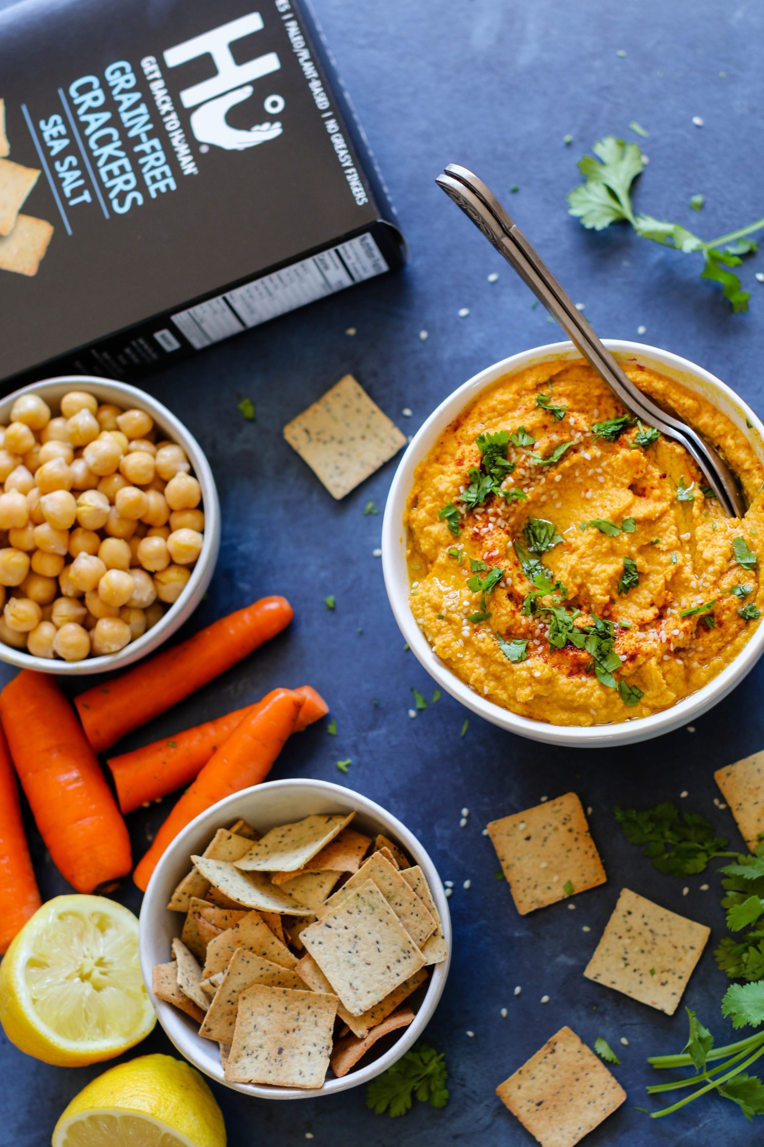 Raw Carrot Hummus served in a bowl with two spoons and fresh herbs surrounded by crackers and carrots and chickpeas with lemon halves and raw carrots