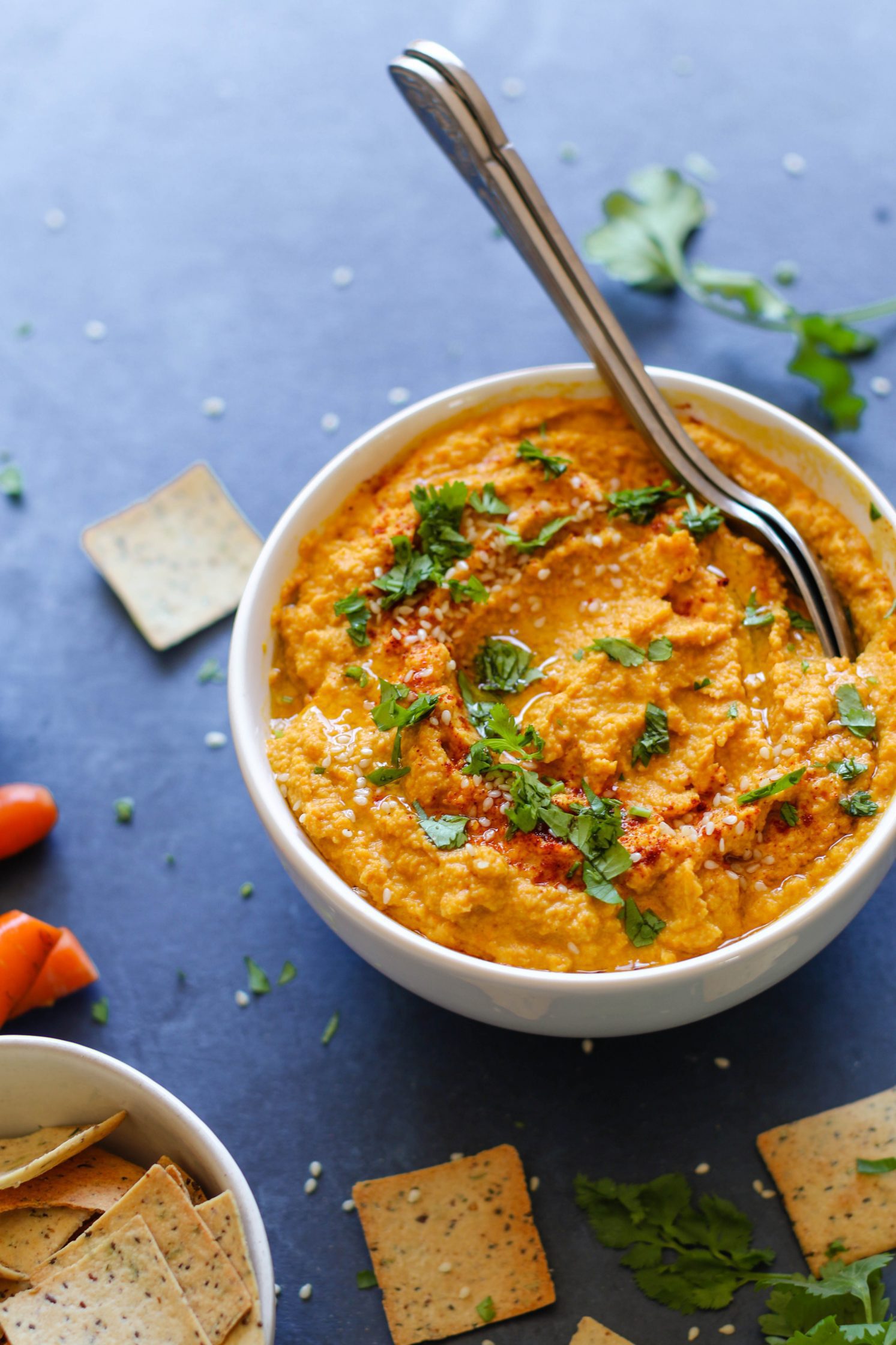 Raw Carrot Hummus served in a bowl with two spoons and fresh herbs surrounded by crackers and carrots and chickpeas with lemon halves and raw carrots