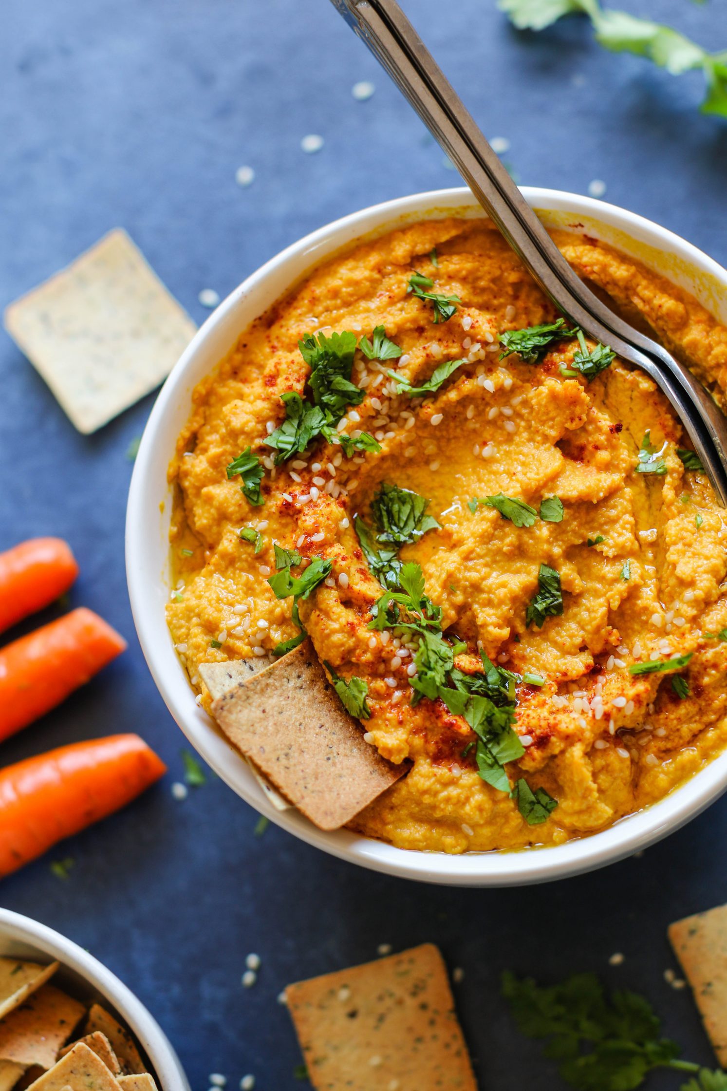 Raw Carrot Hummus served in a bowl with two spoons and fresh herbs surrounded by crackers and carrots and chickpeas with lemon halves and raw carrots
