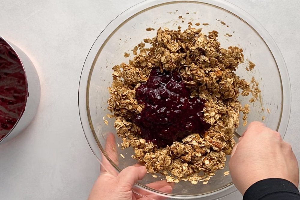 hands mixing berry compote into granola mixture in glass bowl