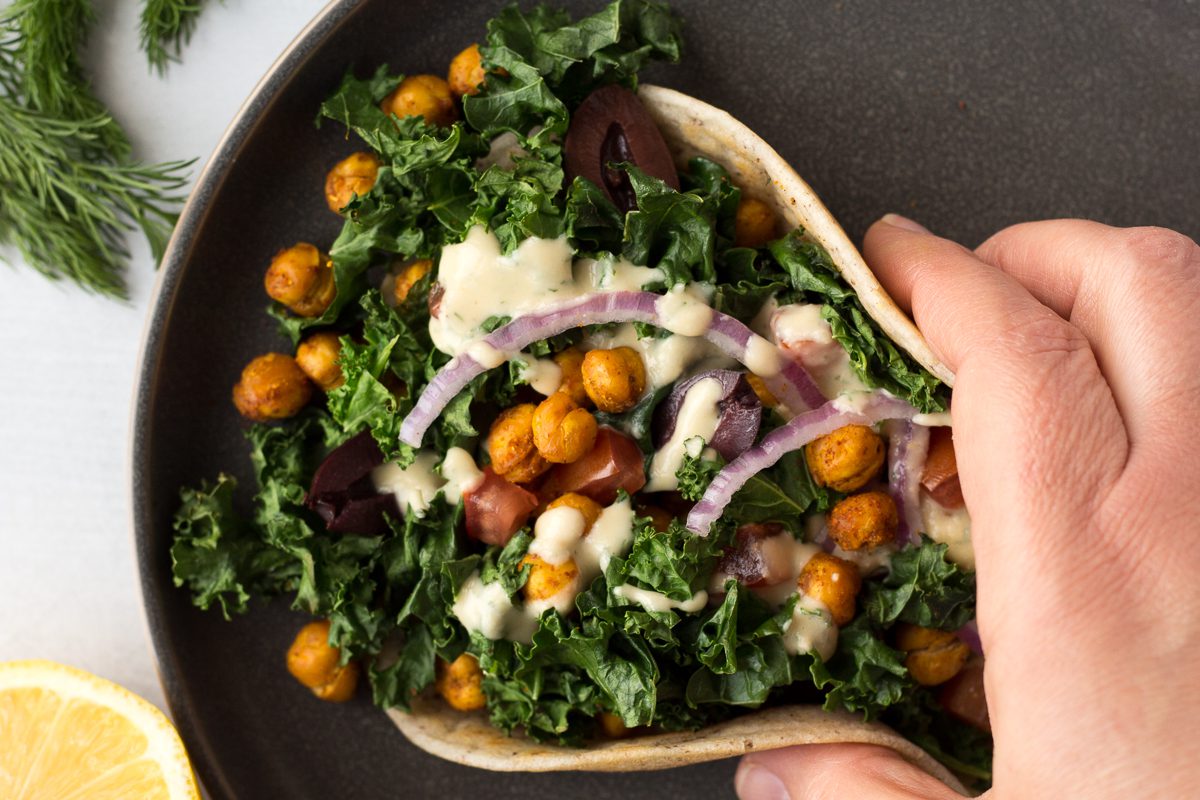 hand holding Grain-Free Flatbread Wraps served on plate with baked kale, chickpeas, red onion, and dill hummus sauce