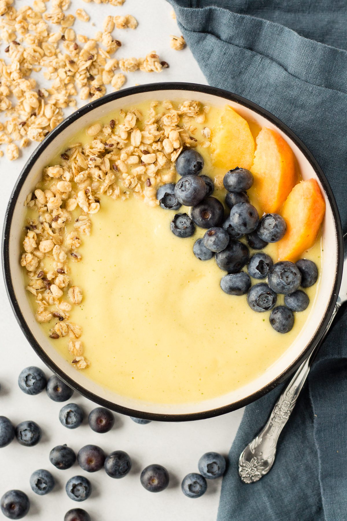 Peach Mango Smoothie Bowl served in bowl topped with blueberries, sliced peaches, and granola with spoon and blue towel 