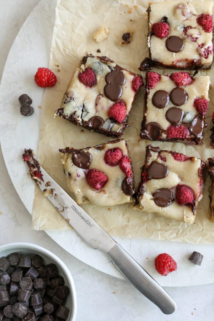 Dark Chocolate Raspberry Blondies cut into squares on parchment paper with raspberries and chocolate chips scattered by Flora & Vino