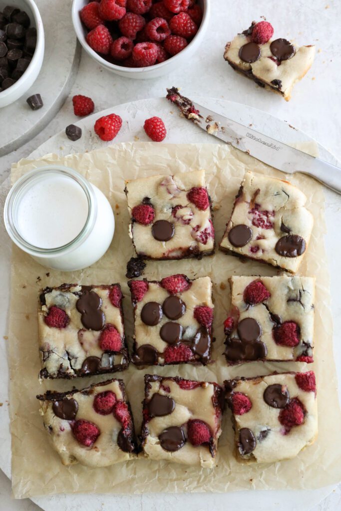 Dark Chocolate Raspberry Blondies cut into squares on parchment paper with raspberries and chocolate chips scattered by Flora & Vino