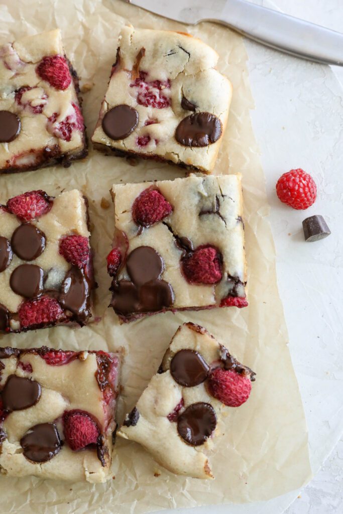 Dark Chocolate Raspberry Blondies cut into squares on parchment paper with raspberries and chocolate chips scattered by Flora & Vino