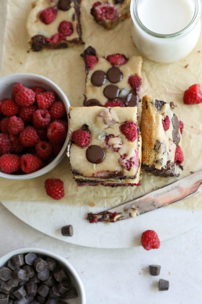 Dark Chocolate Raspberry Blondies stacked with raspberries and dark chocolate scattered and glass of almond milk by Flora & Vino