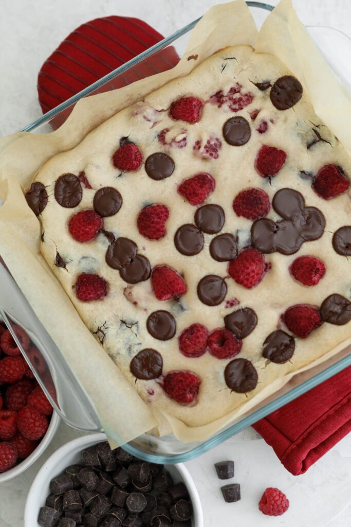 Dark Chocolate Raspberry Blondies baked in parchment lined baking dish on top of red oven mitt by Flora & Vino