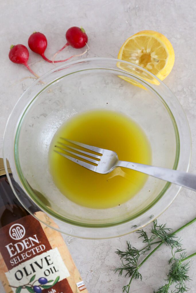 dressing for salad in glass bowl with a fork with radishes, dill, lemon, and Eden Foods Olive Oil on the side