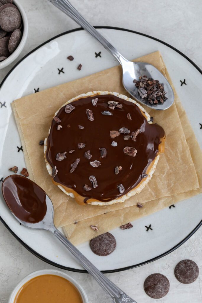 Chocolate Peanut Butter Rice Cake served on parchment paper lined plate with scattered cacao nibs, chocolate wafers, and peanut butter