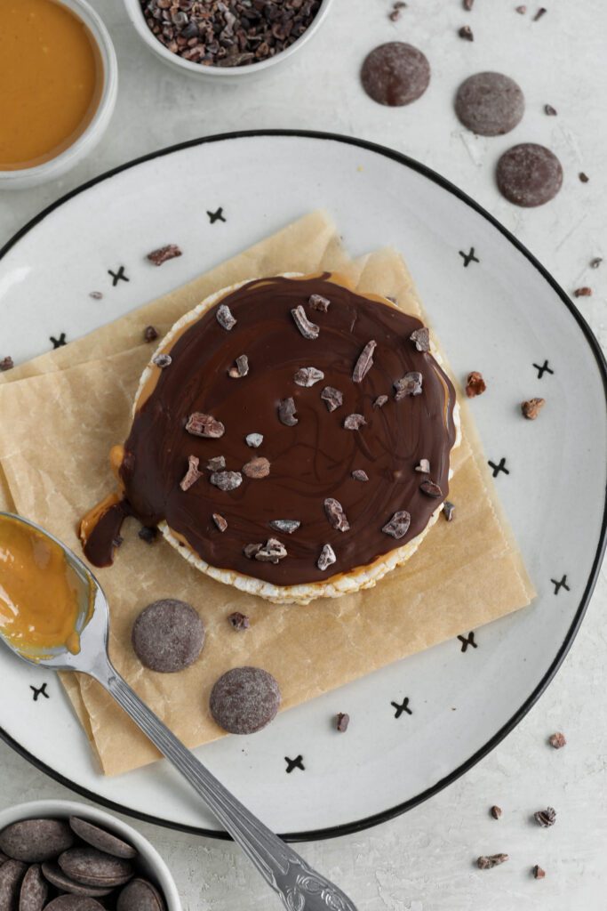 Chocolate Peanut Butter Rice Cake served on parchment paper lined plate with scattered cacao nibs, chocolate wafers, and peanut butter