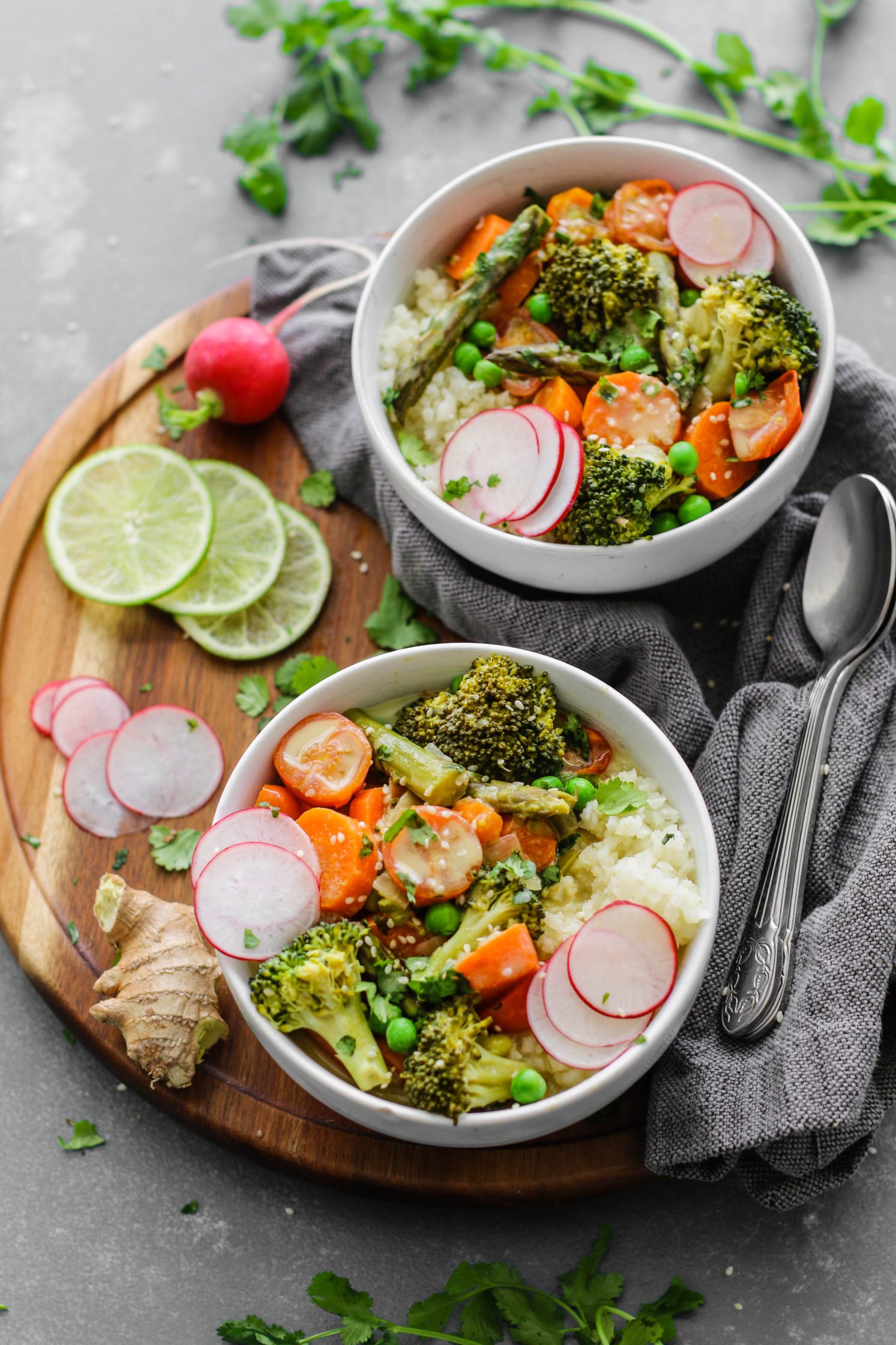 Green Coconut Curry with Cauliflower Rice served on wooden circle board with fresh lime, sliced radish, cilantro, and two spoons on the side