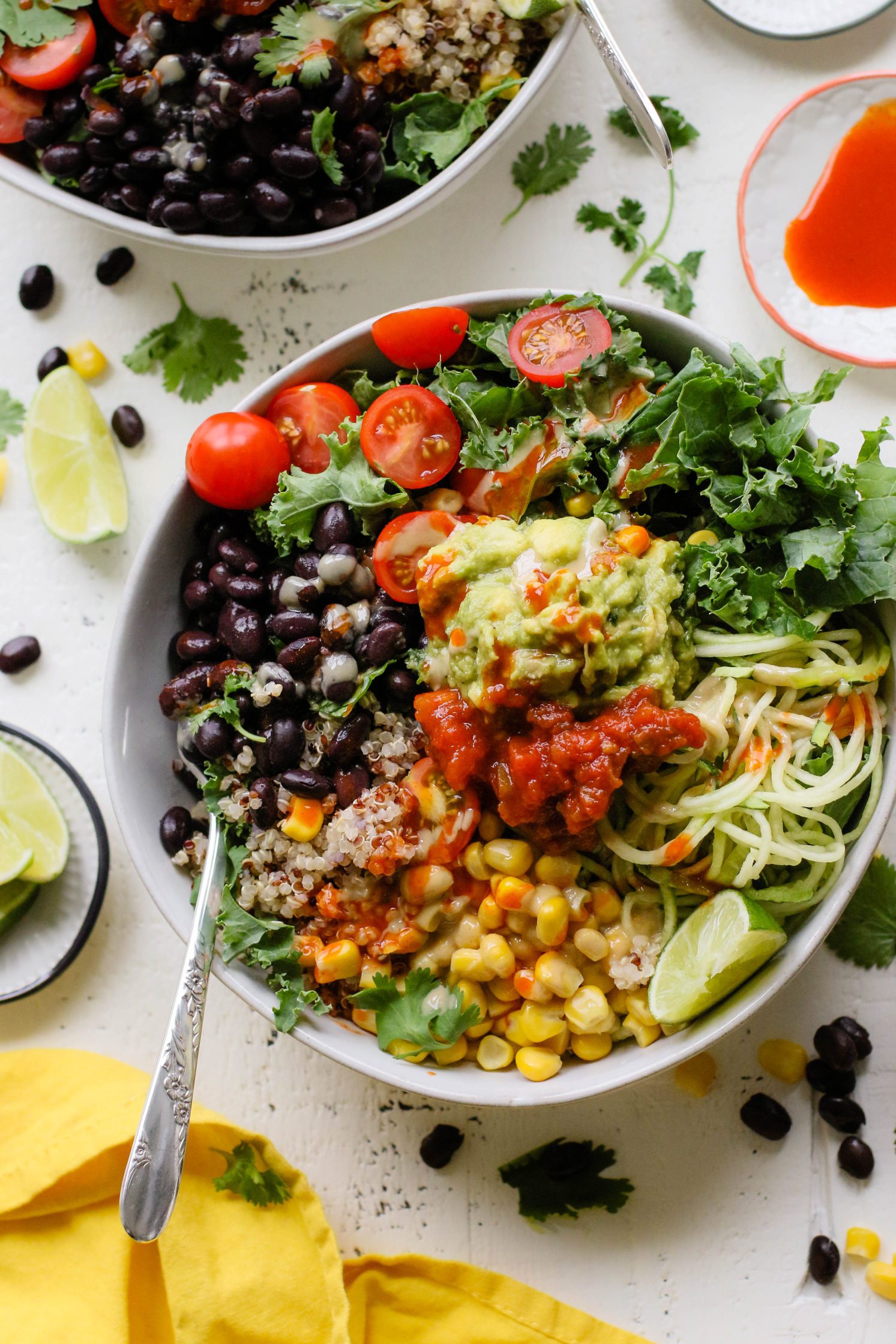 Easy Mexican Macro Bowls served in white bowl with hot sauce, guacamole, corn, cherry tomatoes, kale, zucchini spirals, black beans, and lime with a fork
