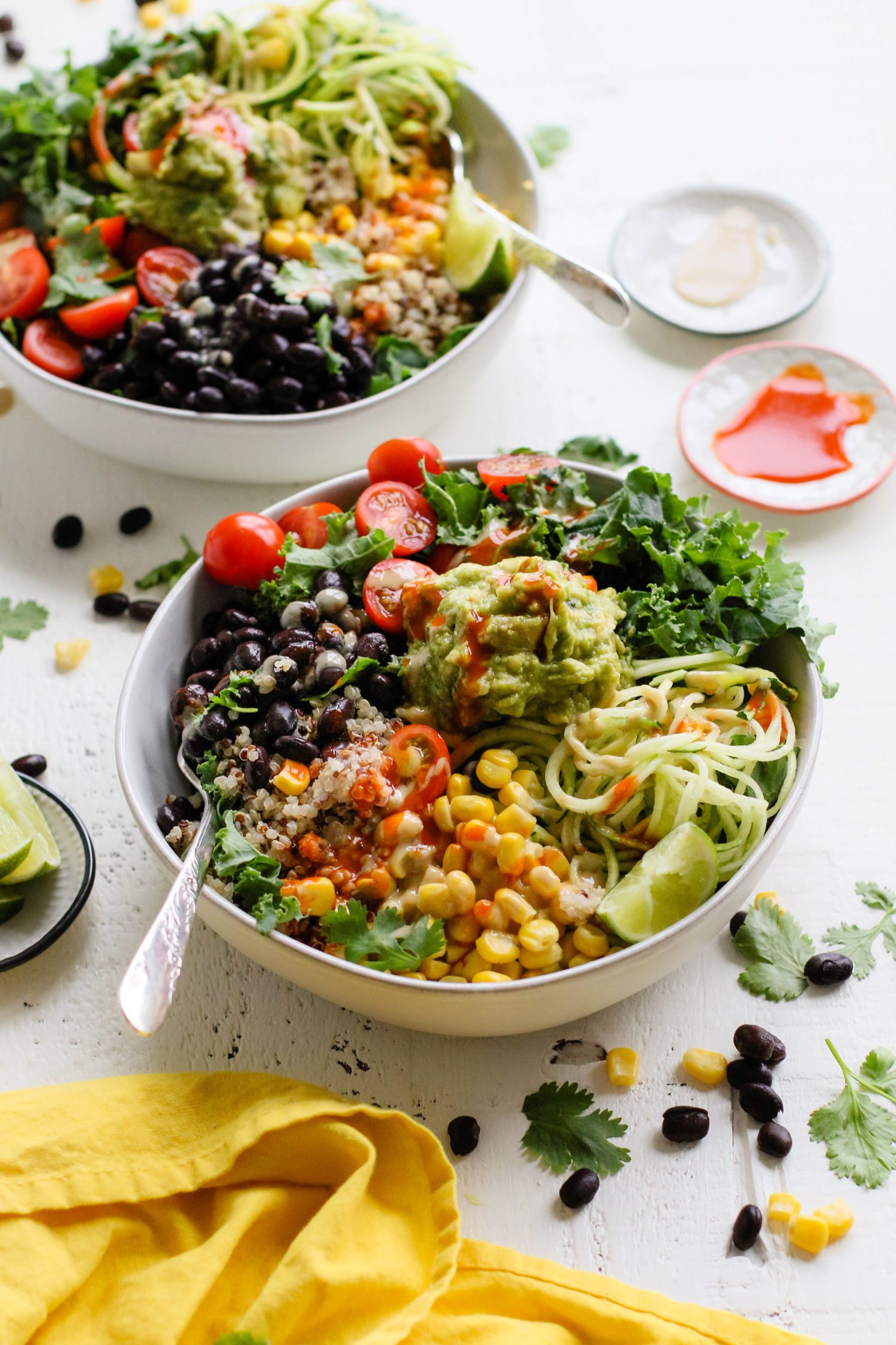 Easy Mexican Macro Bowls served in white bowl with hot sauce, guacamole, corn, cherry tomatoes, kale, zucchini spirals, black beans, and lime with a fork