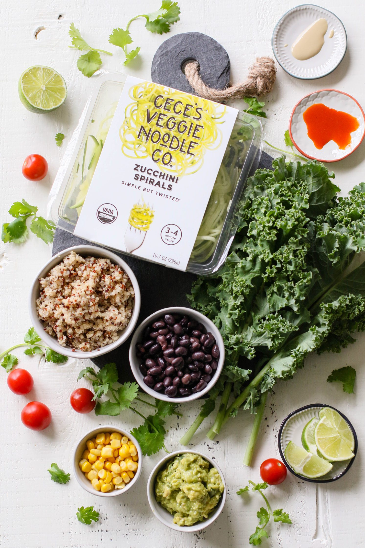 Zucchini spirals in package surrounded by kale, quinoa, black beans, guacamole, corn, lime slices, hot sauce, tahini, and cilantro