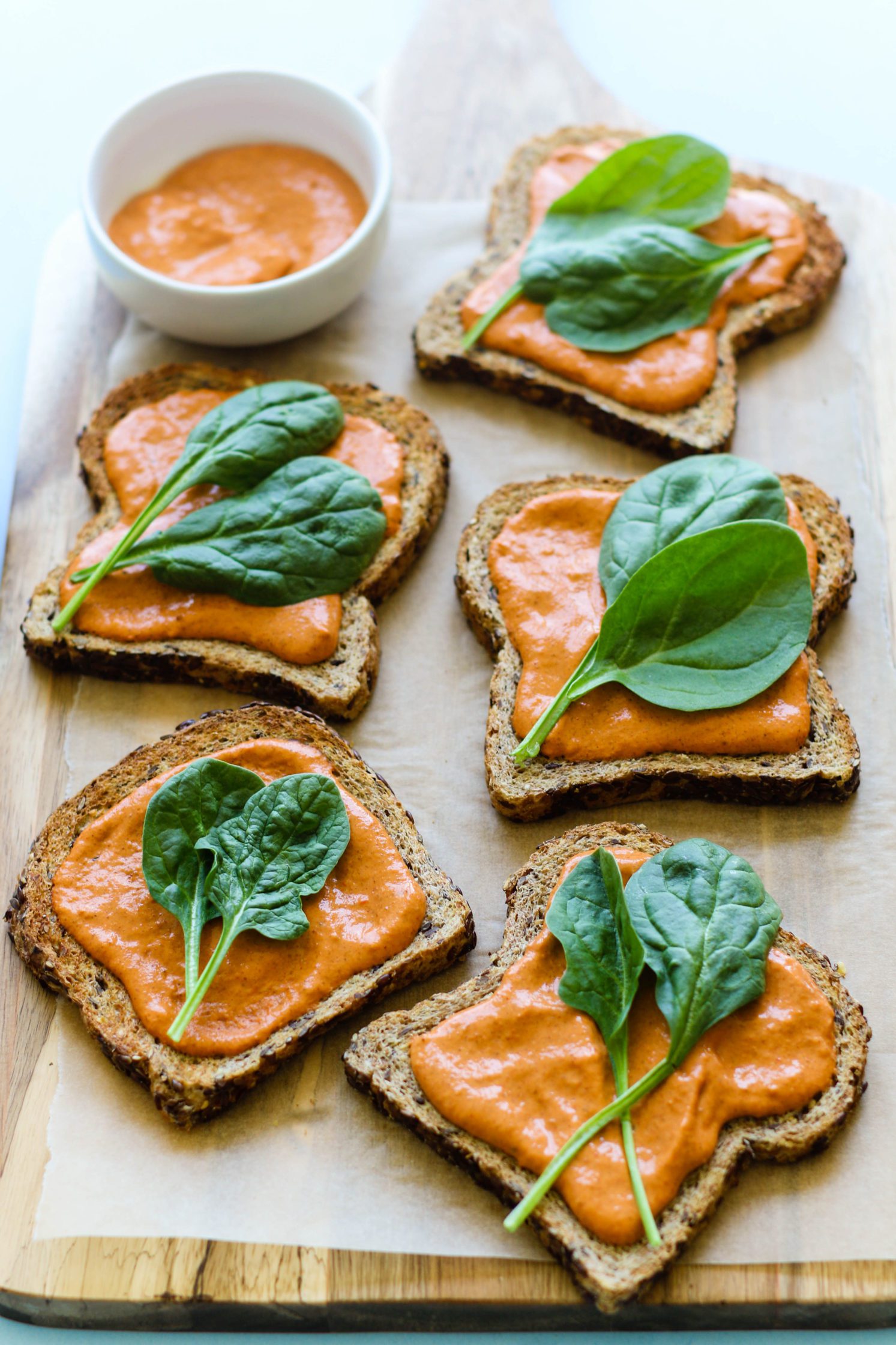 toasted bread topped with romesco sauce and fresh spinach on parchment paper with bowl of romesco sauce 