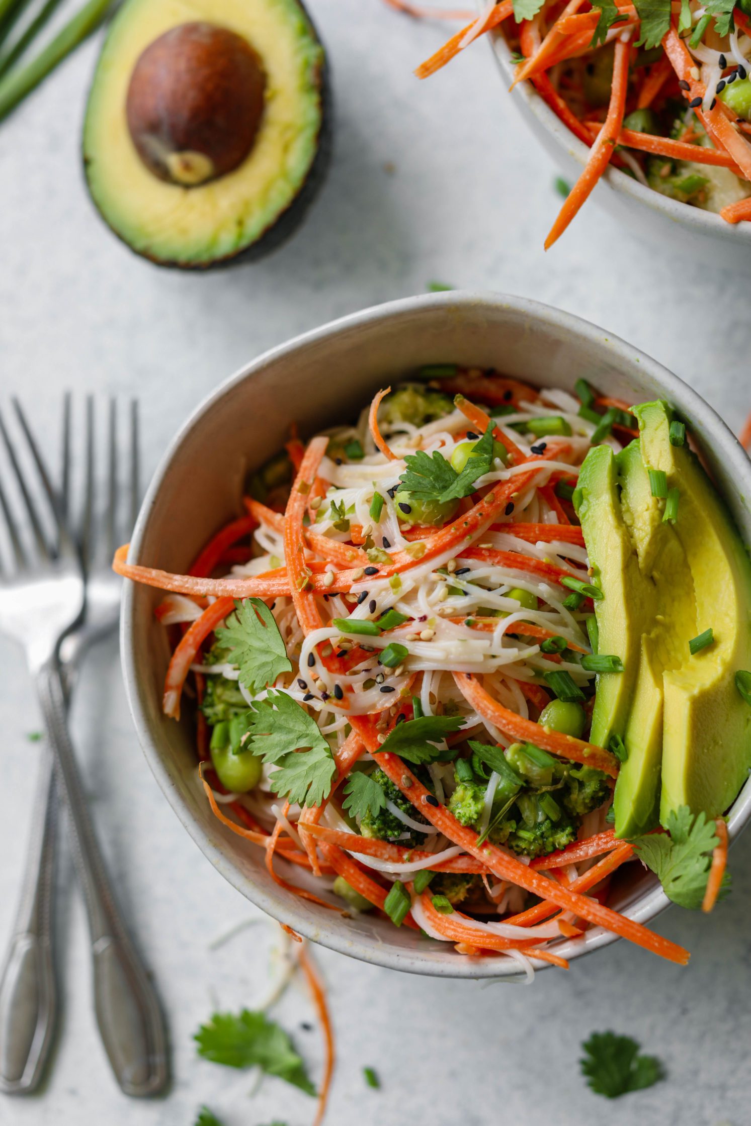 Rainbow Rice Noodle Bowls served in bowl topped with avocado, sesame seeds, and cilantro with two forks