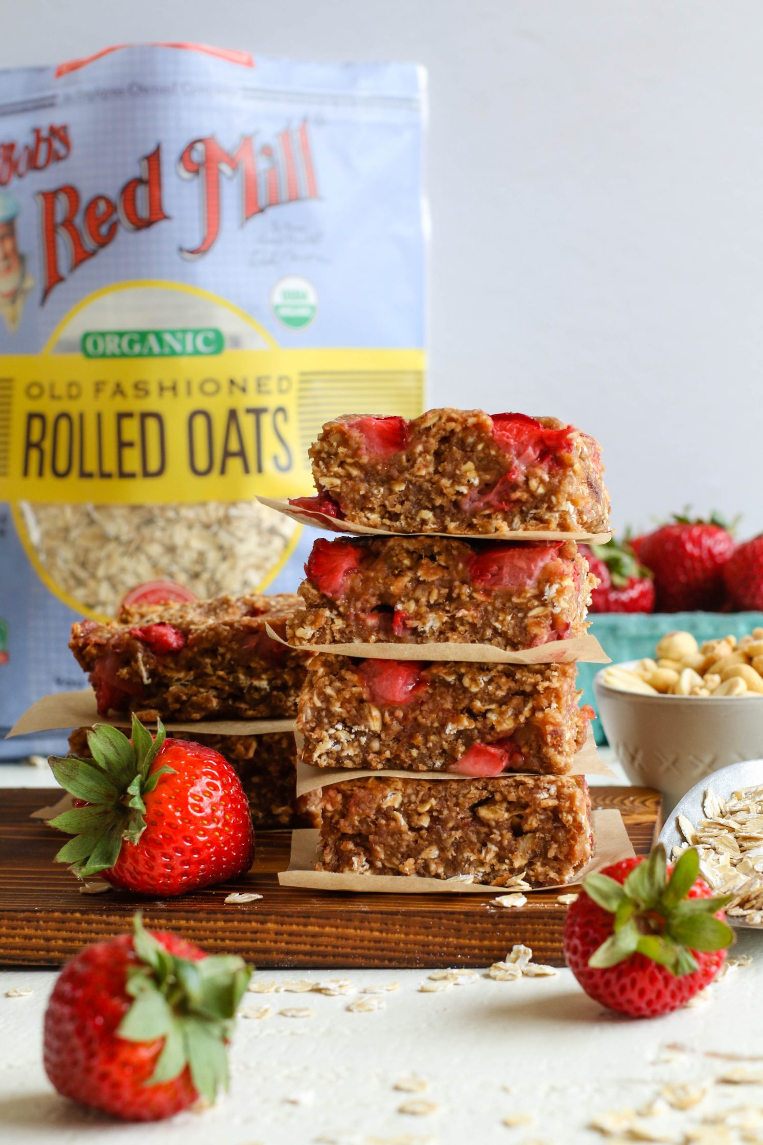 PB&J Baked Breakfast Bars stacked on a parchment lined wooden board with strawberries and scattered oats by Flora & Vino
