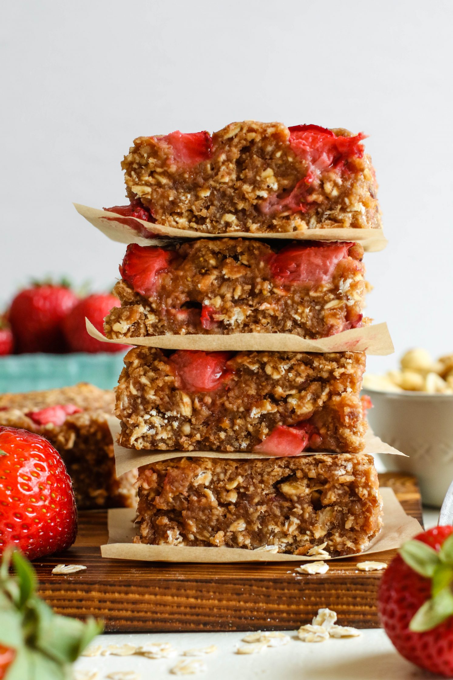 PB&J Baked Breakfast Bars stacked on a parchment lined wooden board with strawberries and scattered oats by Flora & Vino