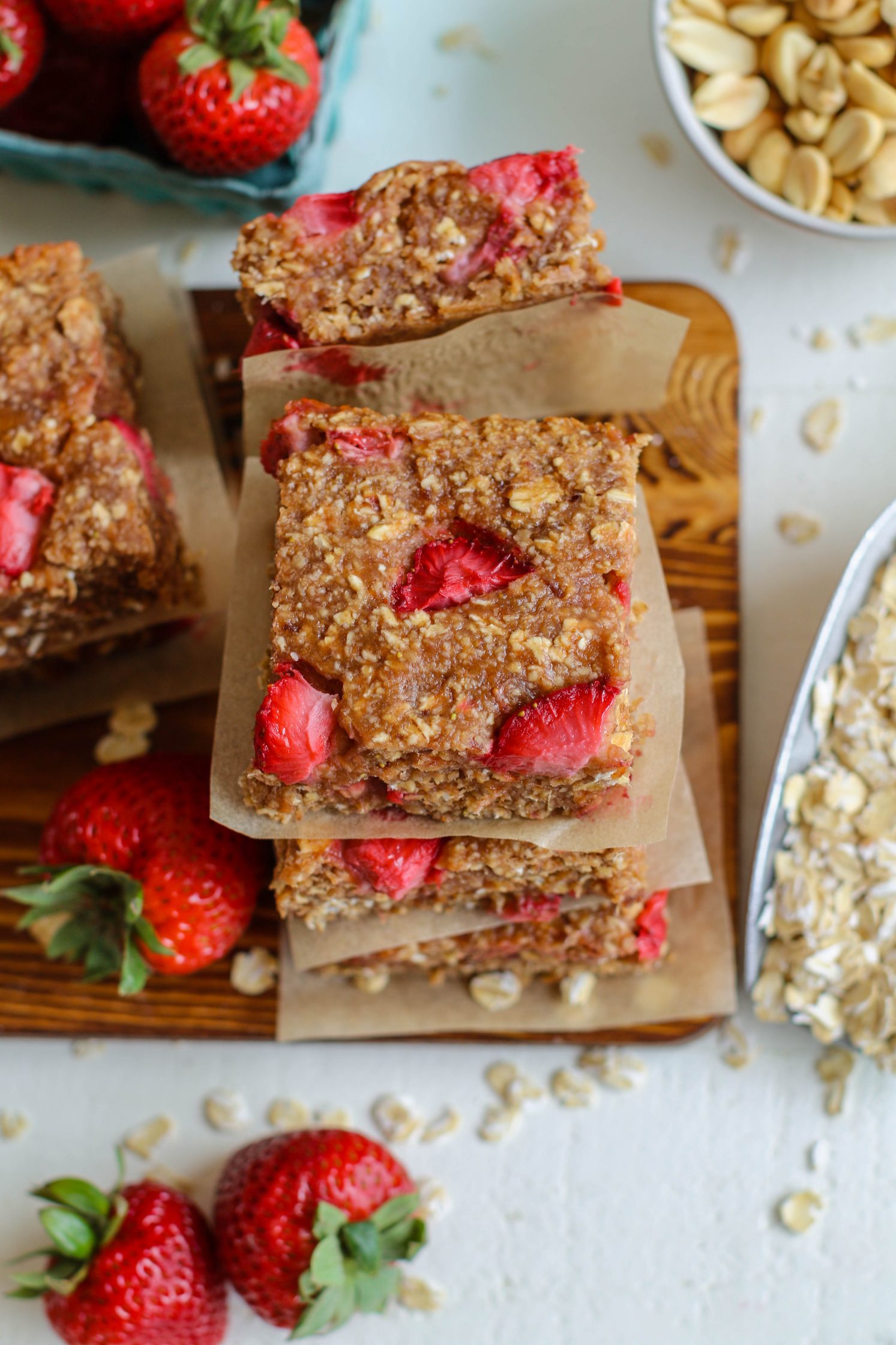 PB&J Baked Breakfast Bars stacked on a parchment lined wooden board with strawberries and scattered oats by Flora & Vino