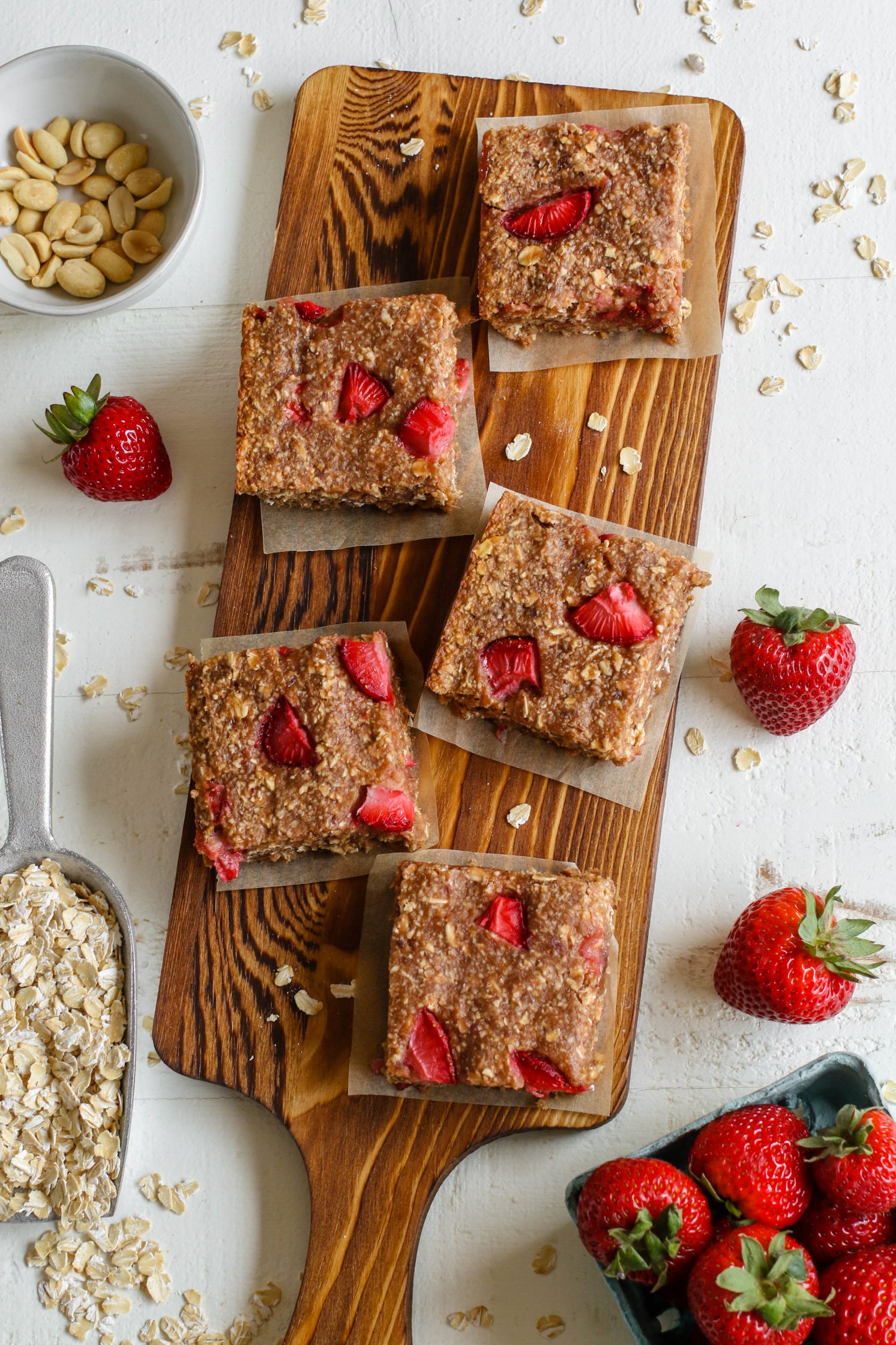 PB&J Baked Breakfast Bars sliced into bars with parchment paper underneath on wooden board with fresh strawberries and peanuts and rolled oats on the side by Flora & Vino