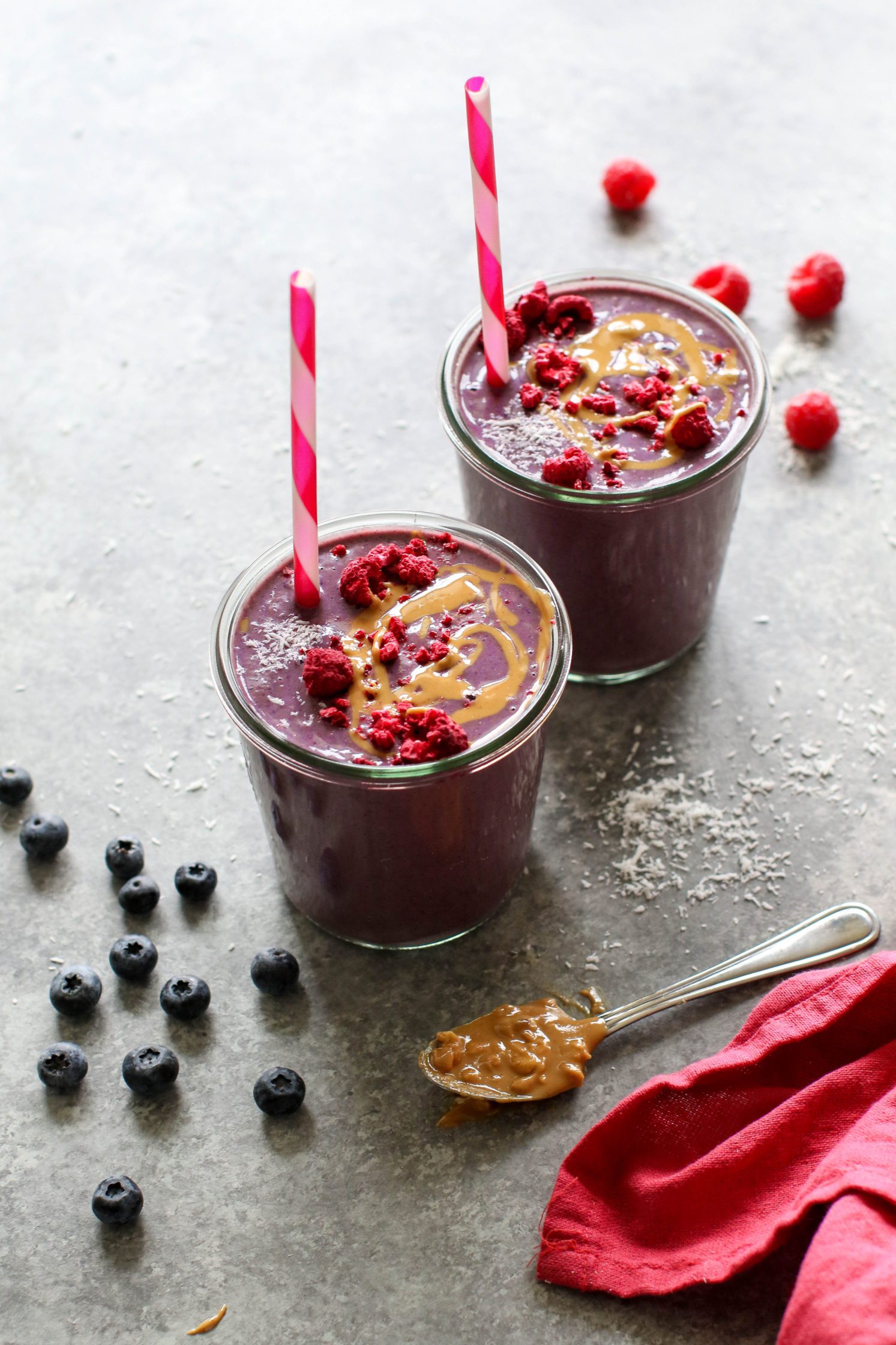 Peanut Butter Jelly & Beet Shakes served in mason jars topped with peanut butter and freeze-dried raspberries with straws. Spoon of peanut butter on the side with a red towel and scattered berries.