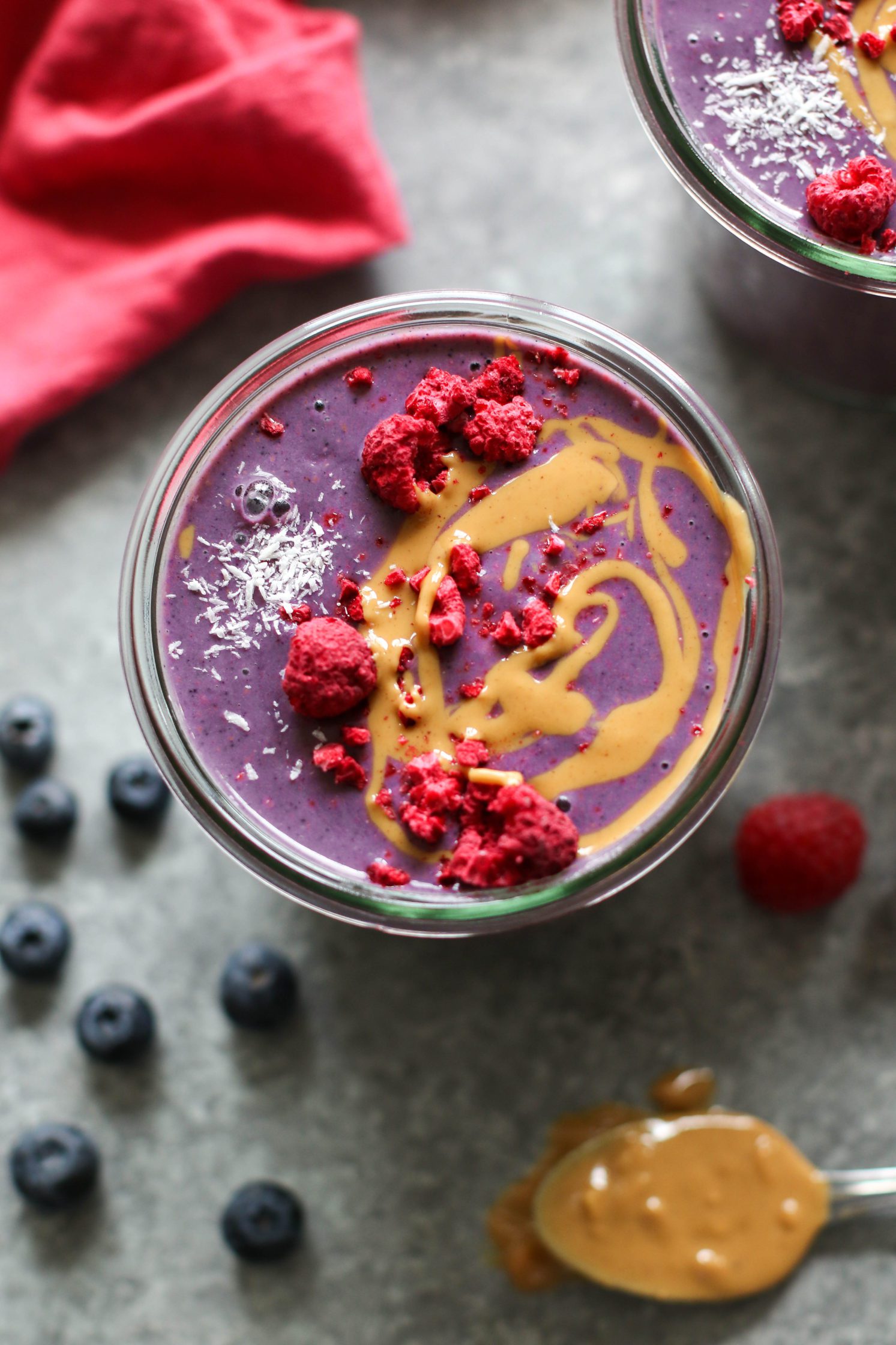 Peanut Butter Jelly & Beet Shakes served in mason jars topped with peanut butter and freeze-dried raspberries with straws. Spoon of peanut butter on the side with a red towel and scattered berries.