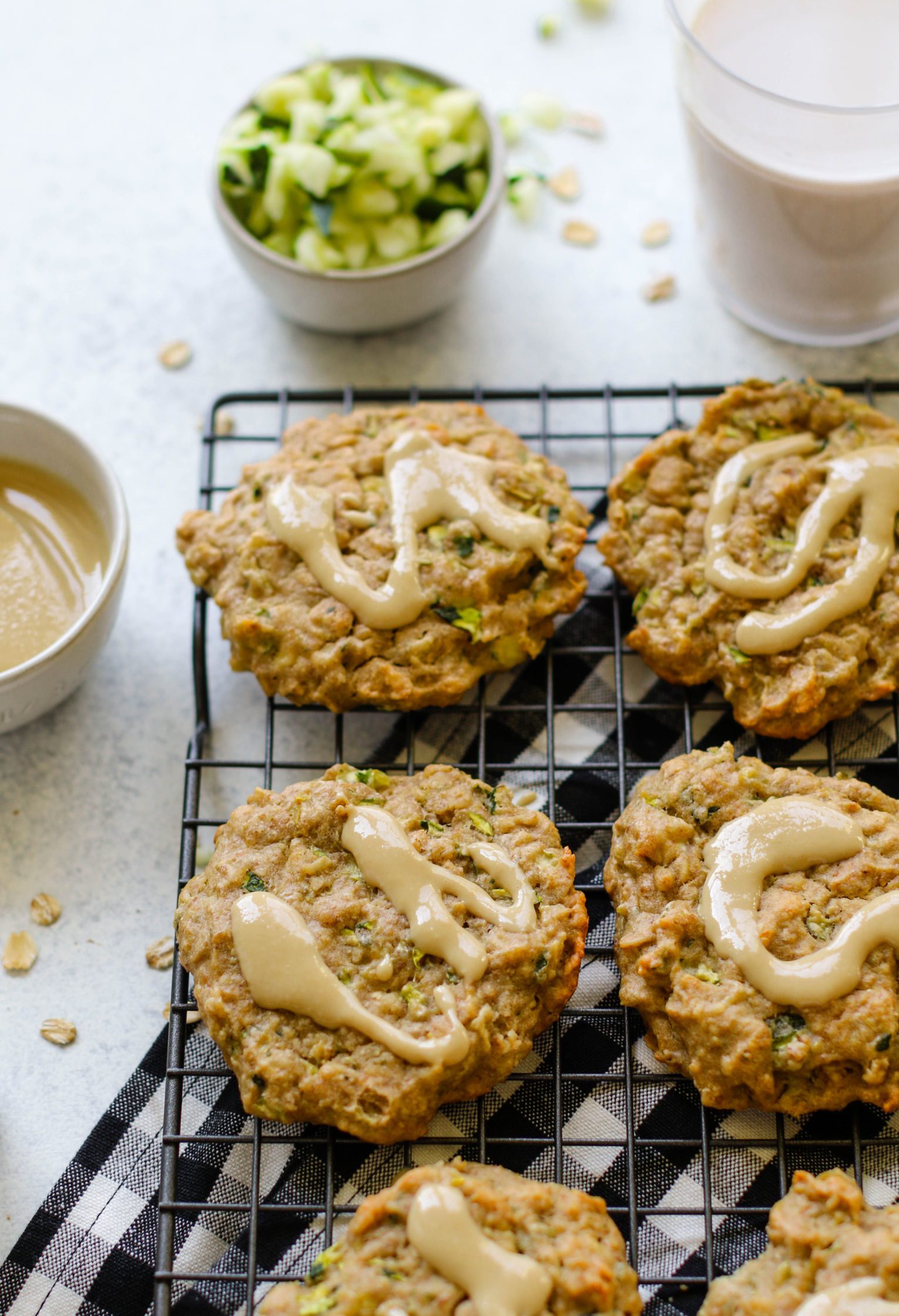 Zucchini Tahini Breakfast Cookies served on cooling rack on top of white and black checkered towel with size of zucchini and almond milk and tahini