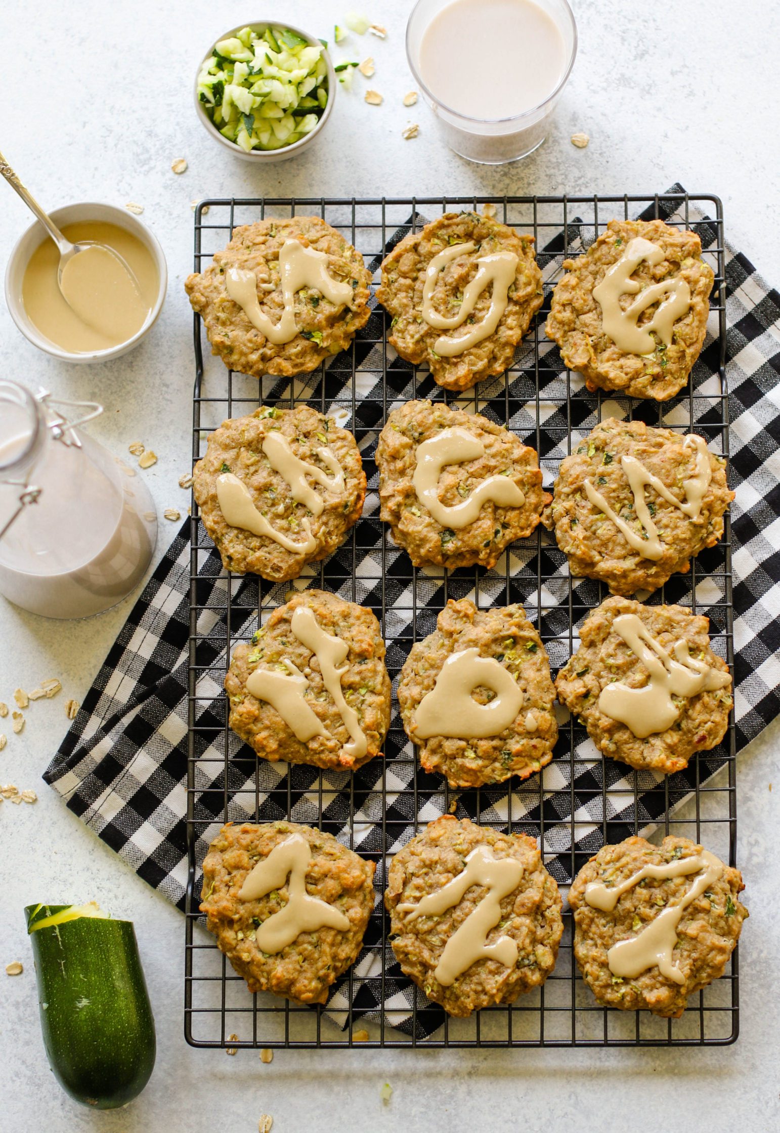 Zucchini Tahini Breakfast Cookies served on cooling rack on top of white and black checkered towel with size of zucchini and almond milk and tahini