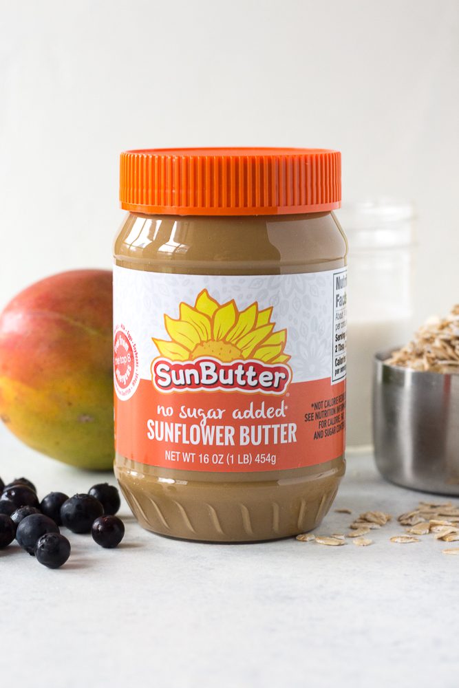 No Sugar Added SunButter jar with scattered blueberries, mango, almond milk, and oats behind it