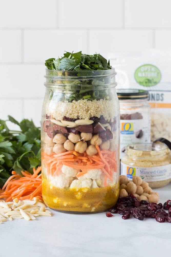 Moroccan Mason Jar Salad in layers surrounded by slivered almonds, shredded carrots, shredded cilantro, chickpeas, and cranberries