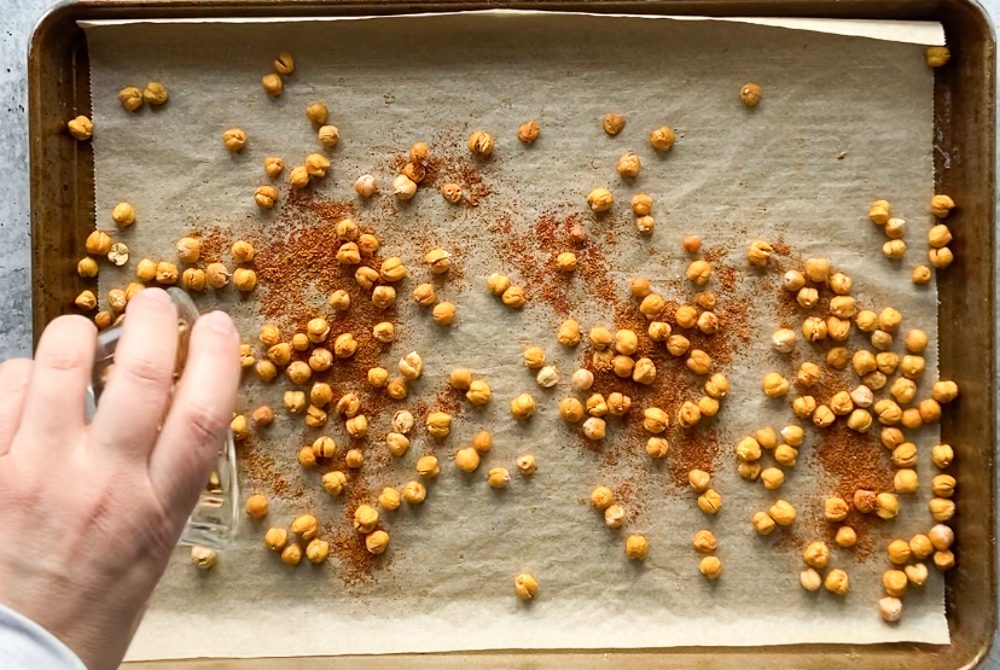 adding spices to roasted chickpeas on parchment lined baking sheet by Flora & Vino