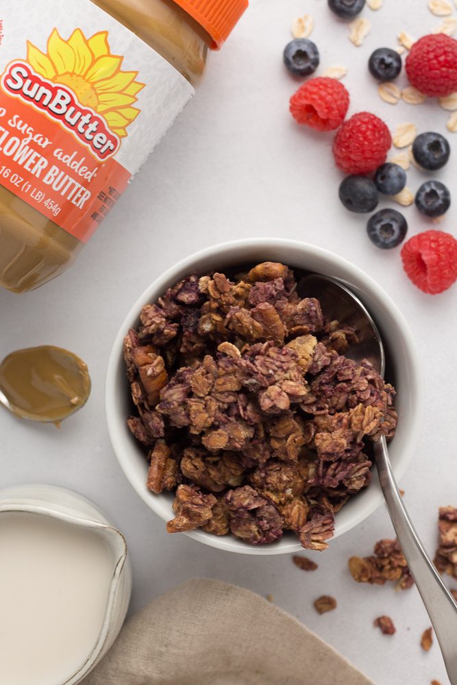 Triple Berry Granola served in bowl with spoon on side and raspberries and blueberries behind and granola scattered