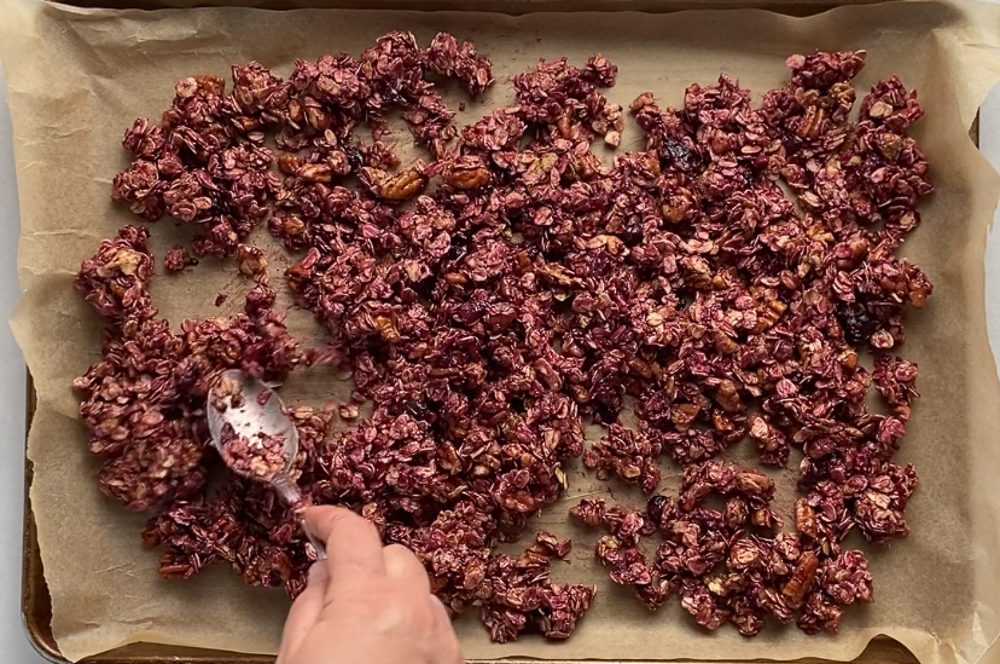 hand using spoon to spread Triple Berry Granola on parchment lined baking sheet