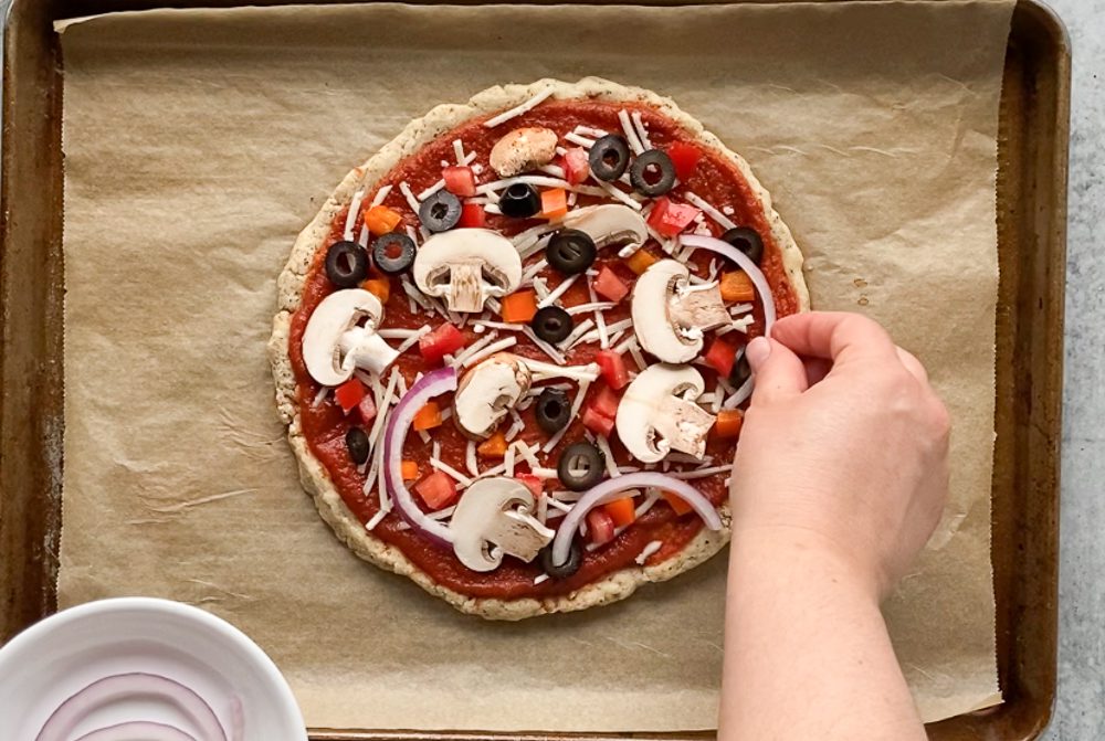 hand adding vegetable ingredients to pizza on parchment lined baking sheet 